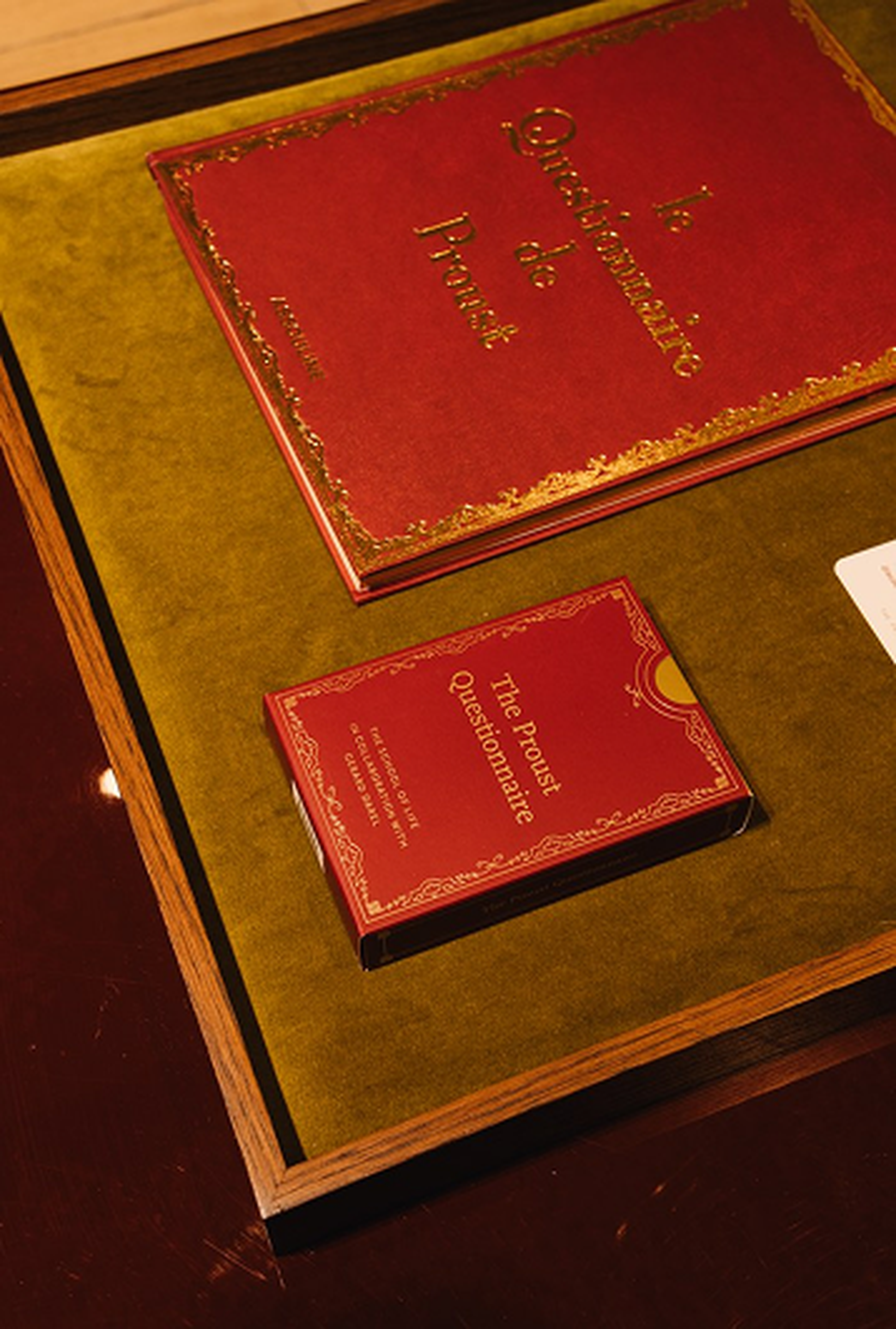 A red book titled "Le Questionnaire de Proust" and a smaller book "The Proust Questionnaire" are displayed on a green surface in a wooden frame.