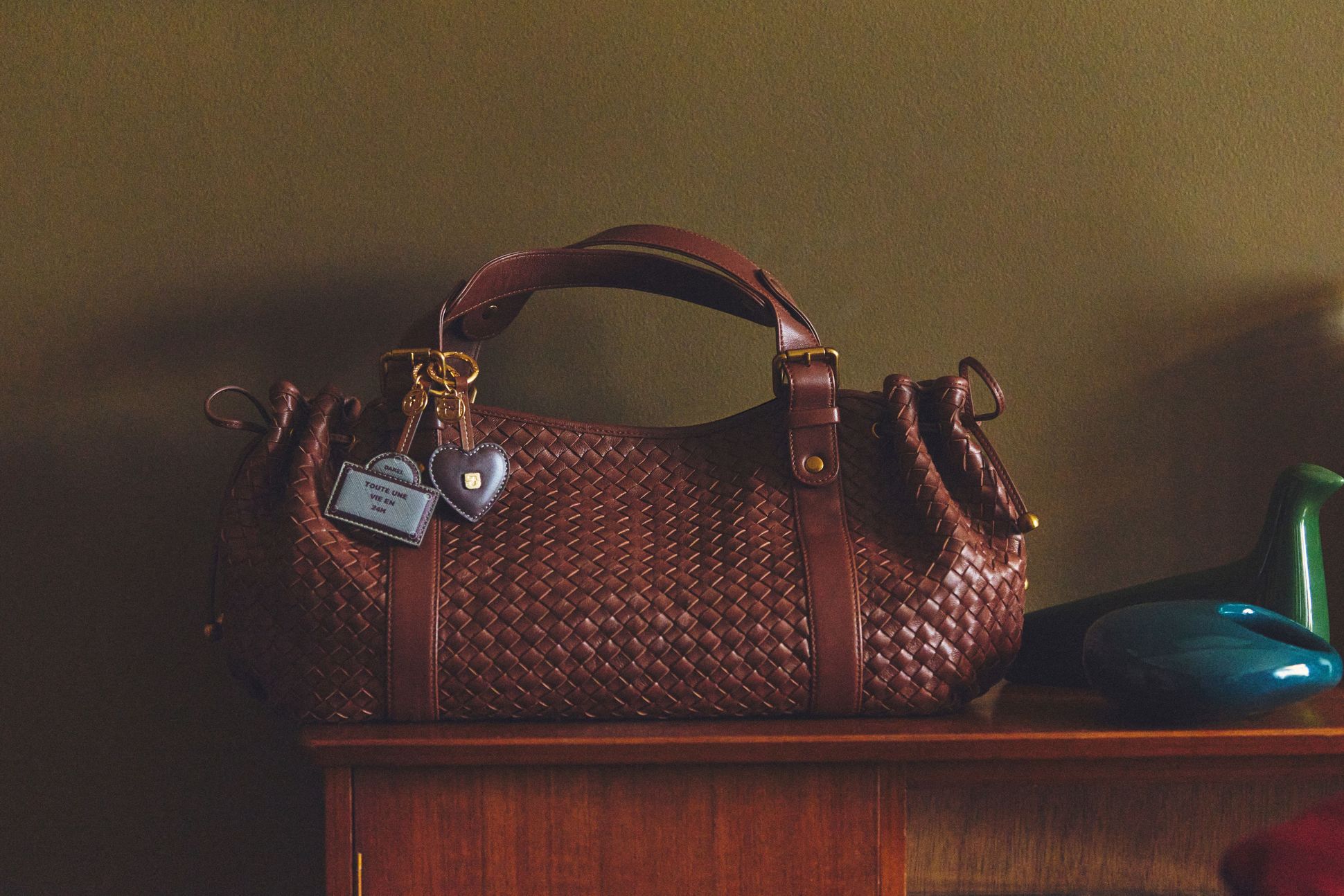 Brown woven leather handbag on a wooden surface, adorned with a blue tag and heart-shaped keychain.