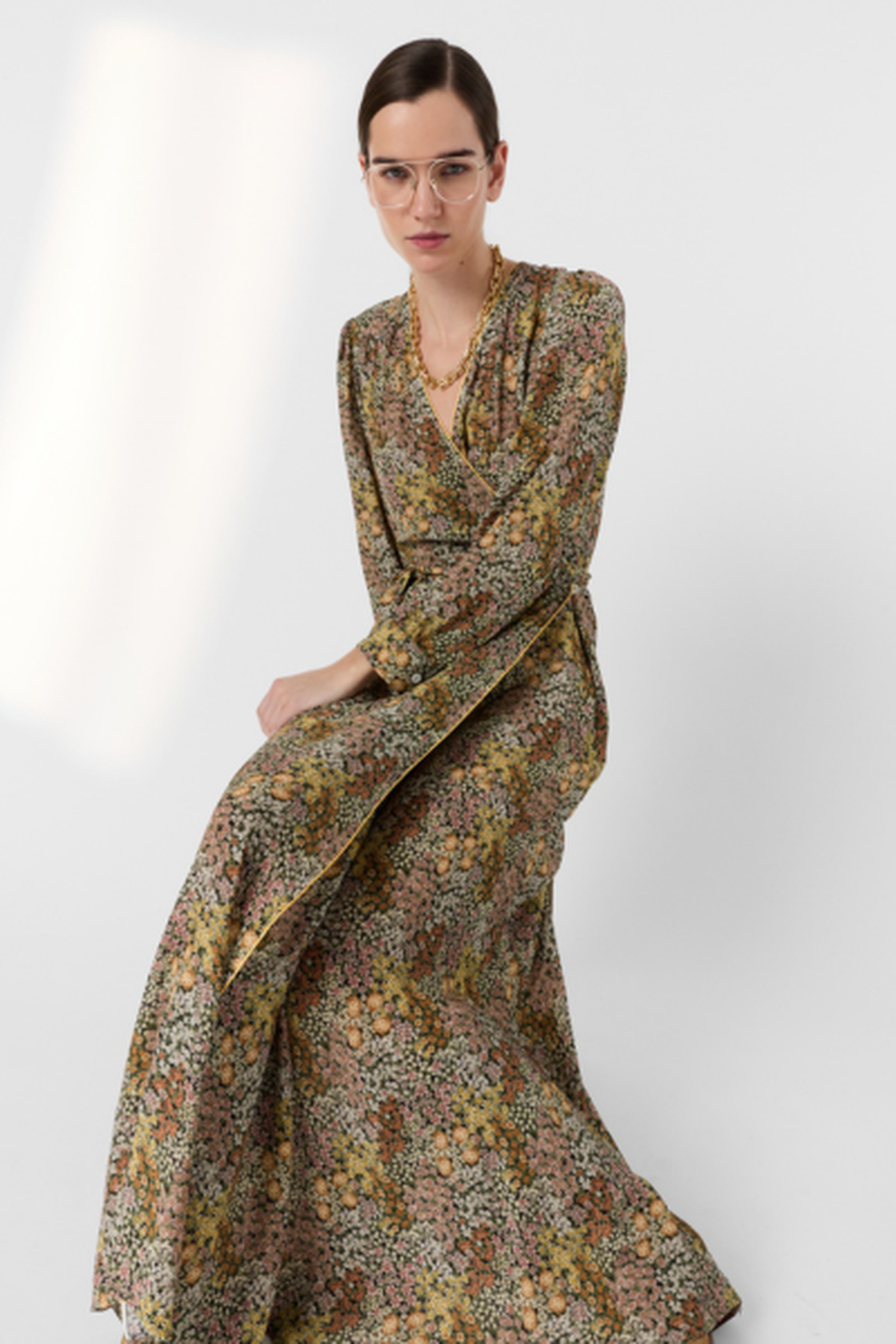 Person seated, wearing glasses and an elegant floral long dress against a plain background.
