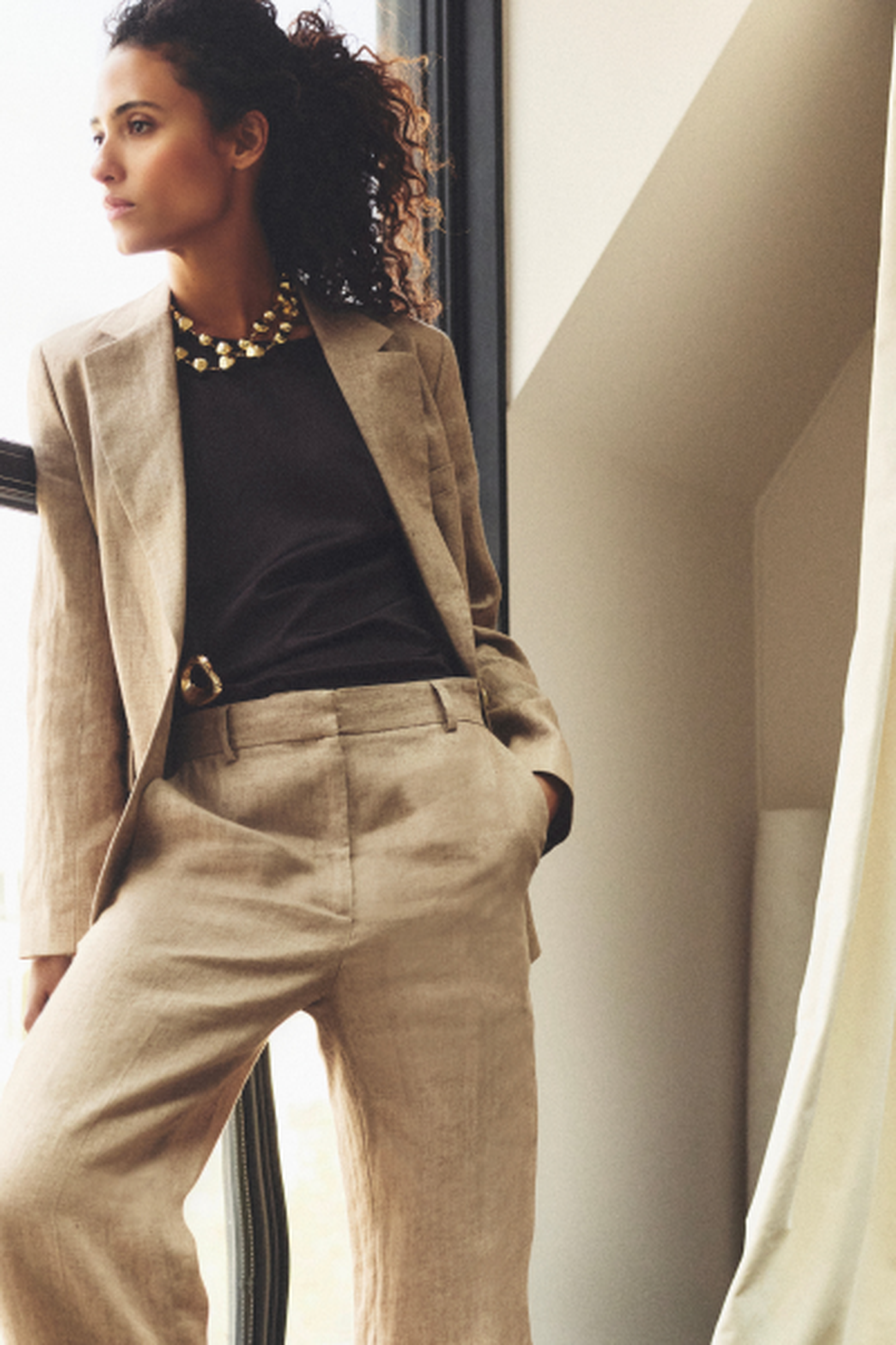 Woman in a beige suit and black top, wearing a chunky necklace, gazing out a window, standing in a modern, softly lit room.