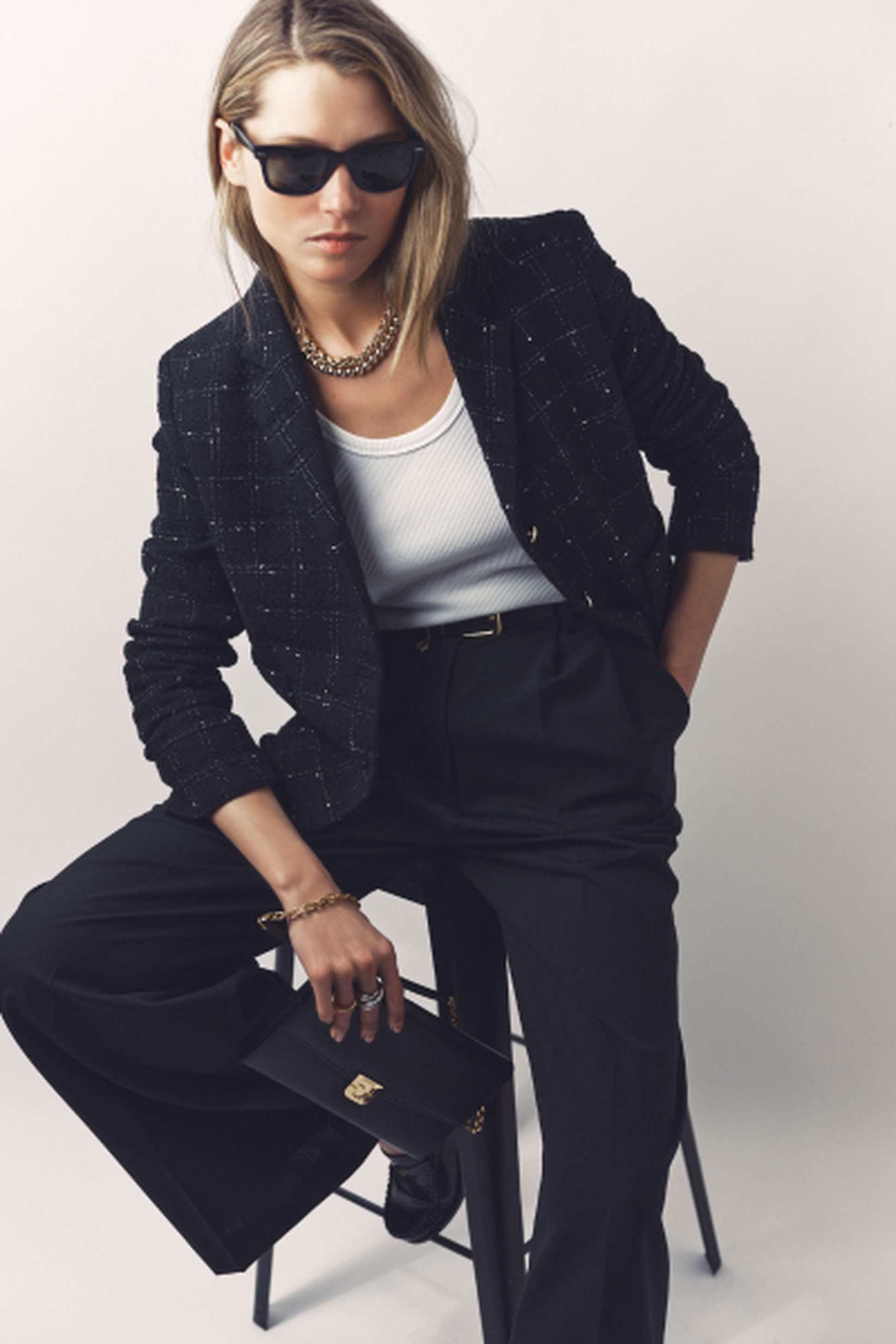 Person in a stylish black blazer and pants, white top, and sunglasses, sitting on a stool. They hold a small clutch and wear a chain necklace.