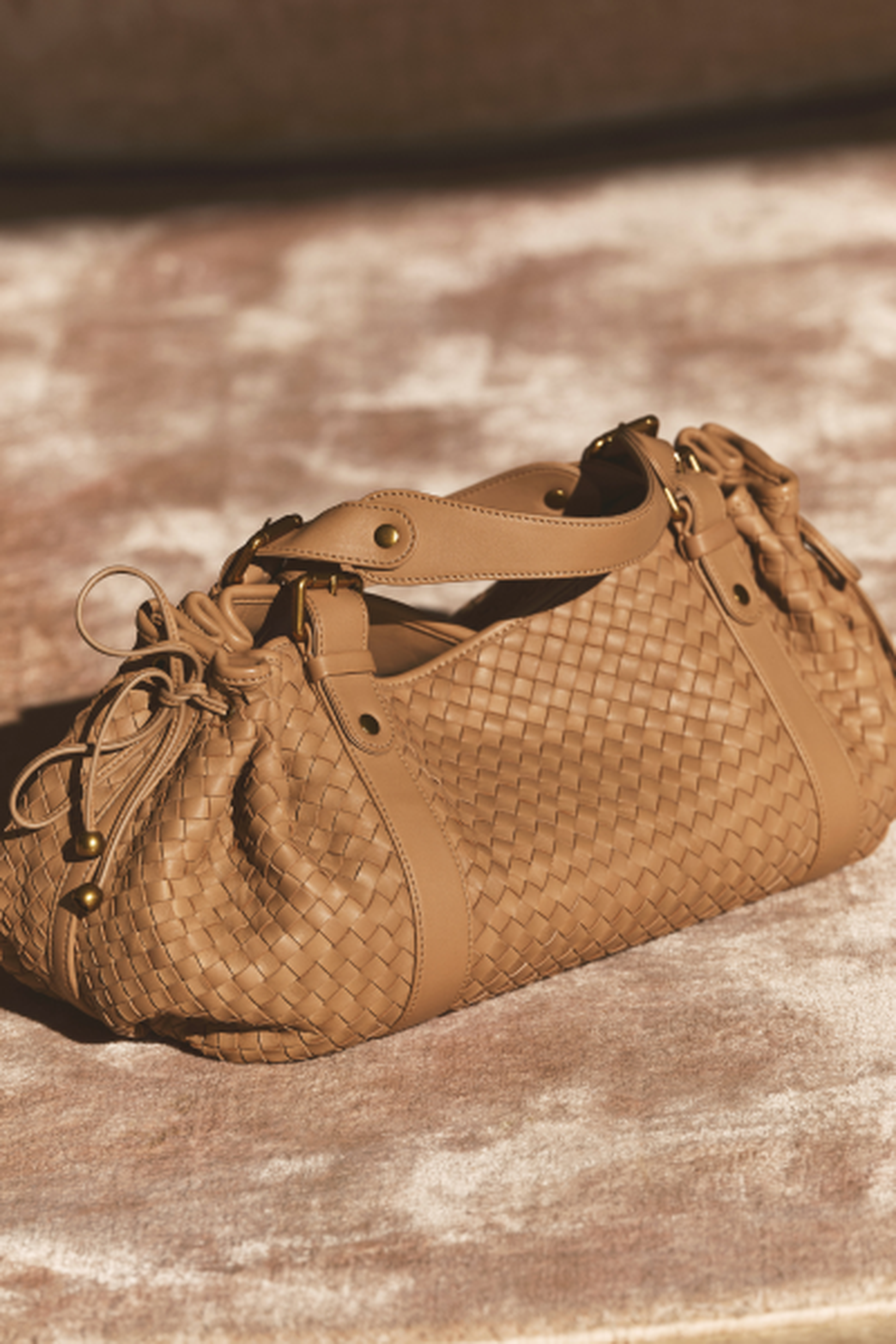 Tan woven leather handbag with drawstring details and metal accents, placed on a plush beige surface.