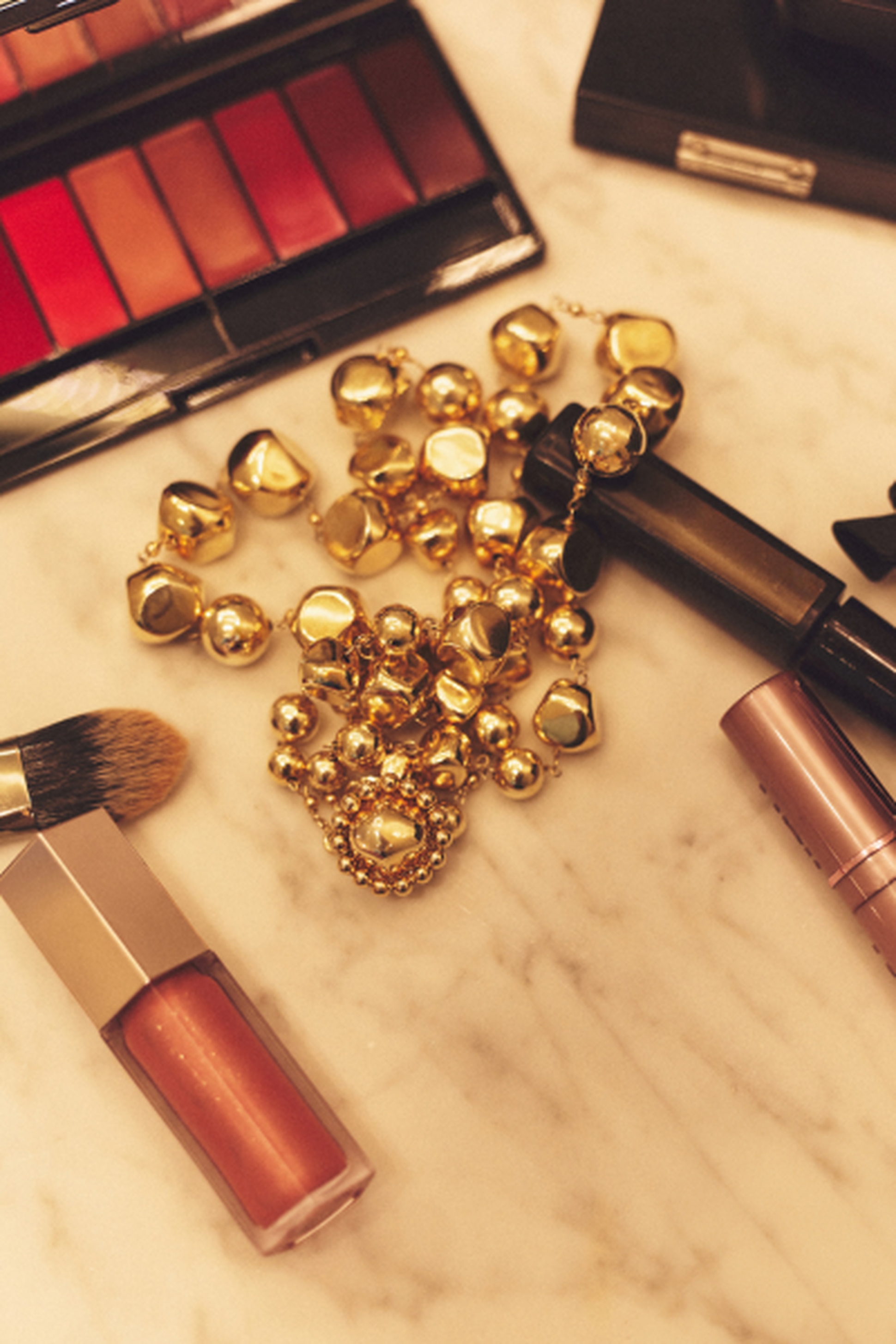 Makeup items, gold jewelry, and brushes arranged on a marble surface, featuring a lipstick palette and lip gloss.