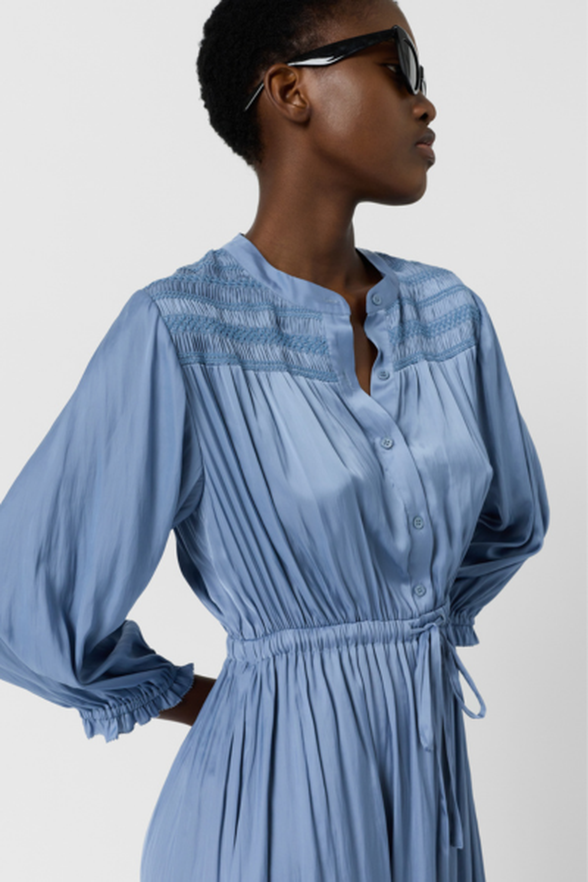 Person wearing a blue smocked dress with long sleeves and a tie waist, against a plain background.