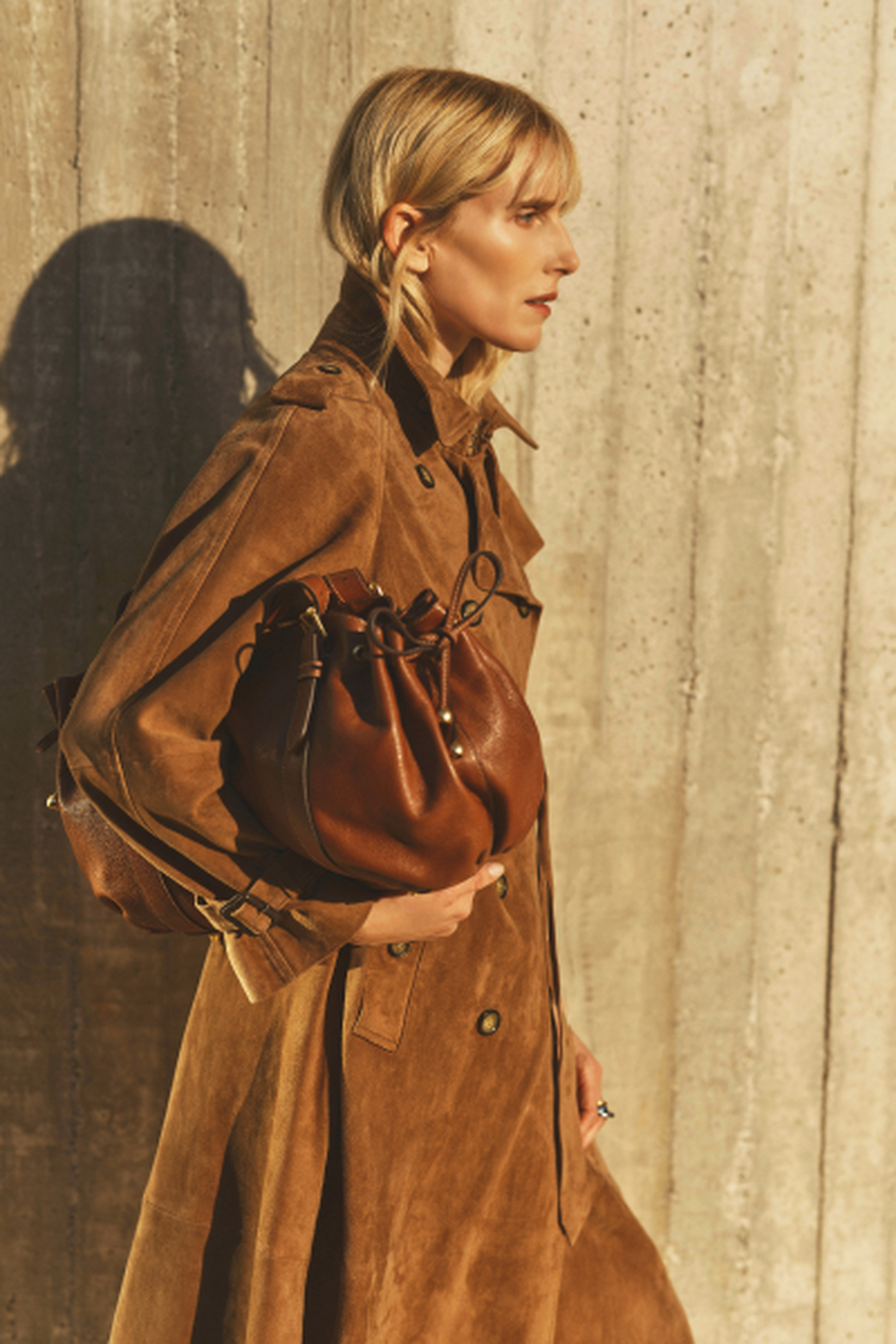 A woman in a brown trench coat holds a leather bag, standing against a textured concrete wall in warm sunlight.
