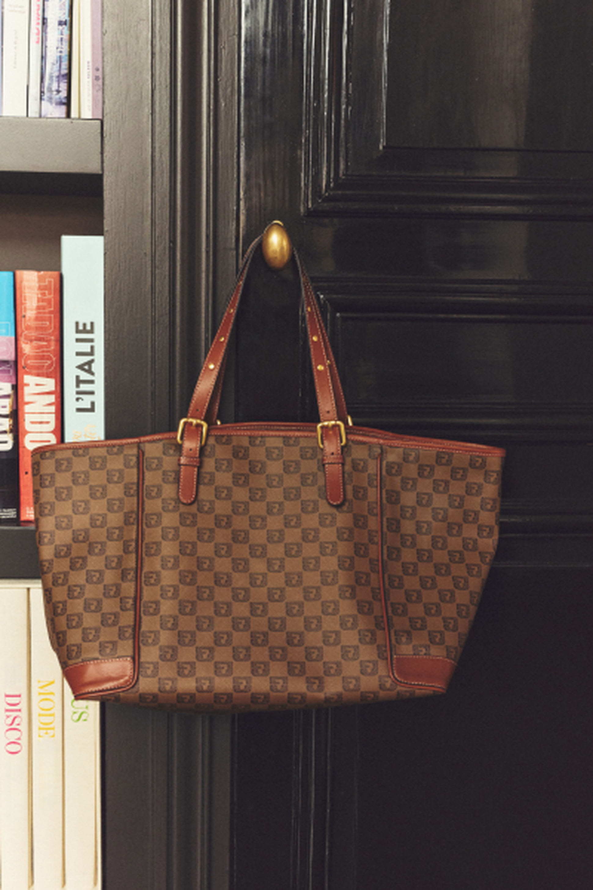 A brown patterned tote bag with leather straps hanging on a black door knob next to a bookshelf with various books.