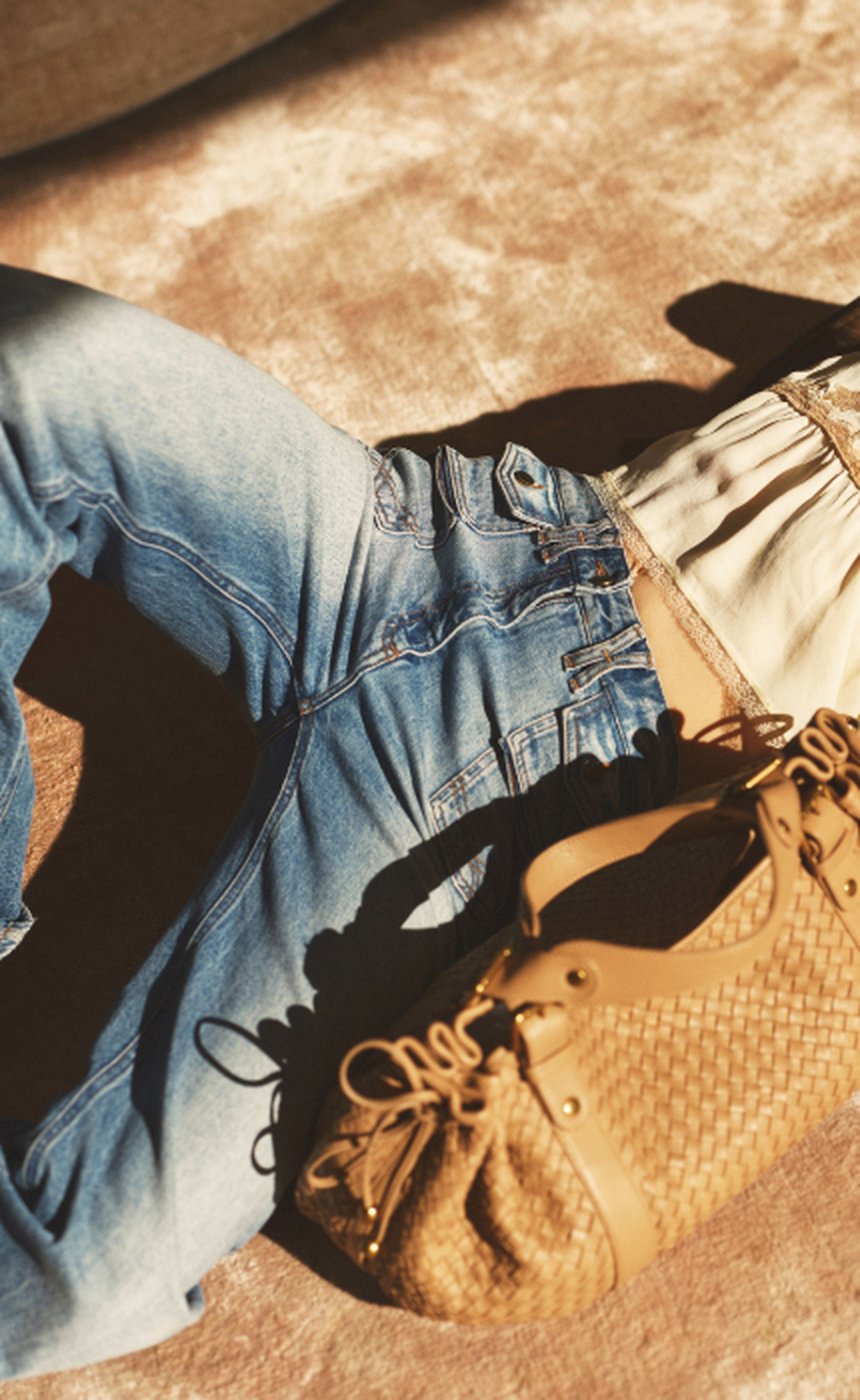 Person lying on a rug, wearing blue jeans and a white top, with a woven tan handbag beside them. Sunlight casts soft shadows.
