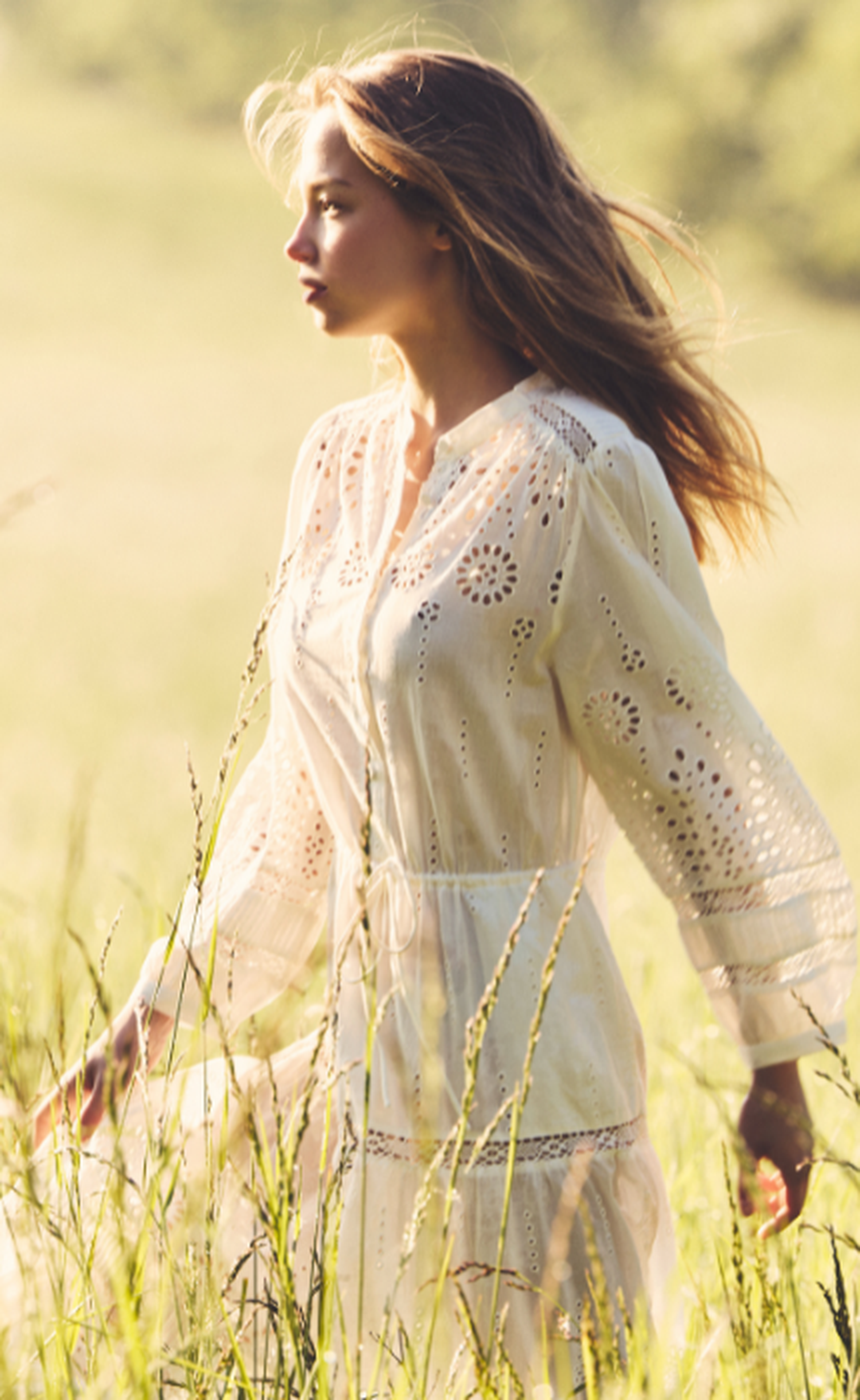 Woman in a white lace dress walking through a sunlit field, with long hair flowing and a serene expression.