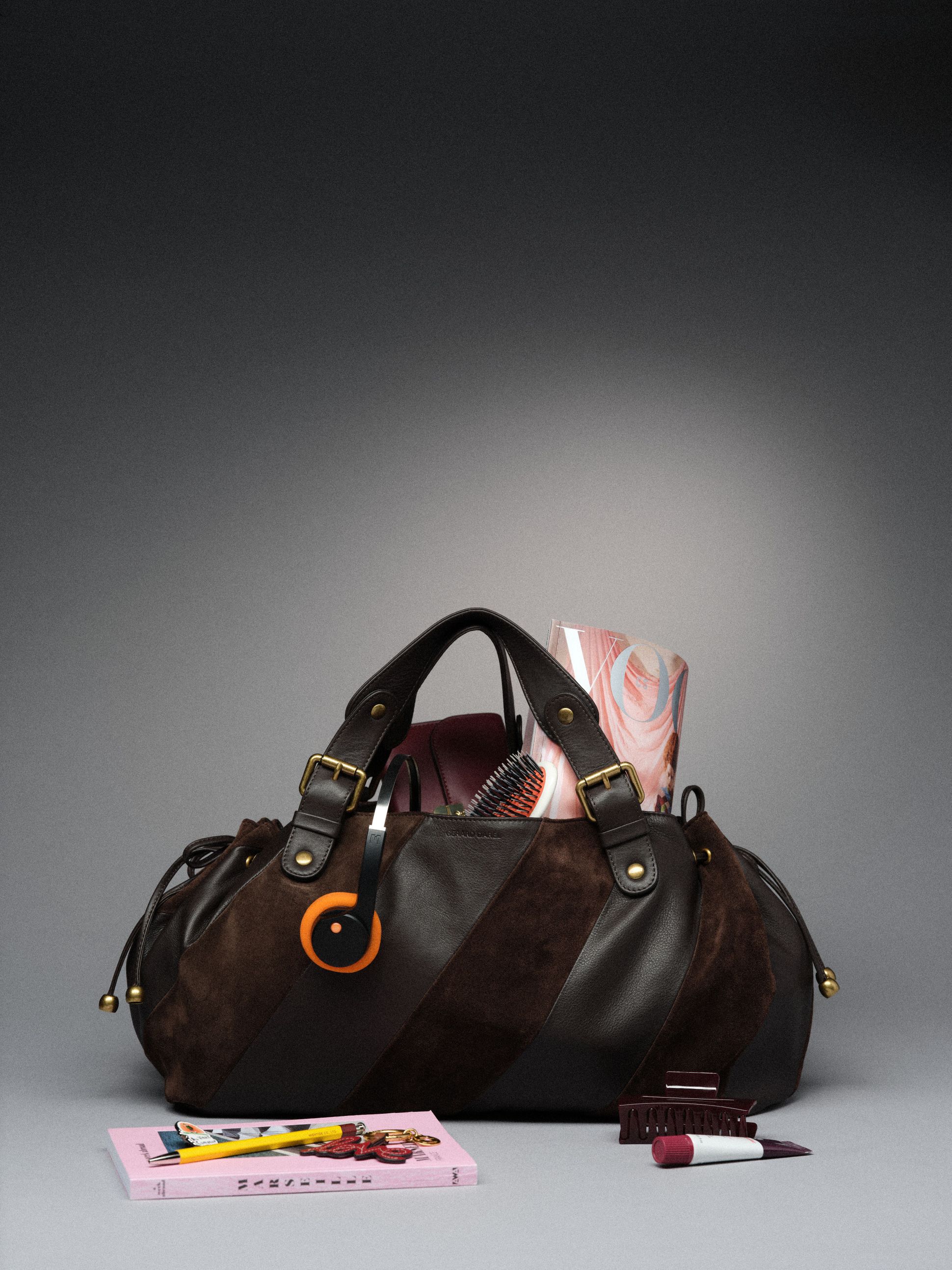A stylish brown bag with gold accents sits on a gray surface, surrounded by a book, pen, lip balm, hair clip, and a brush peeking from inside.