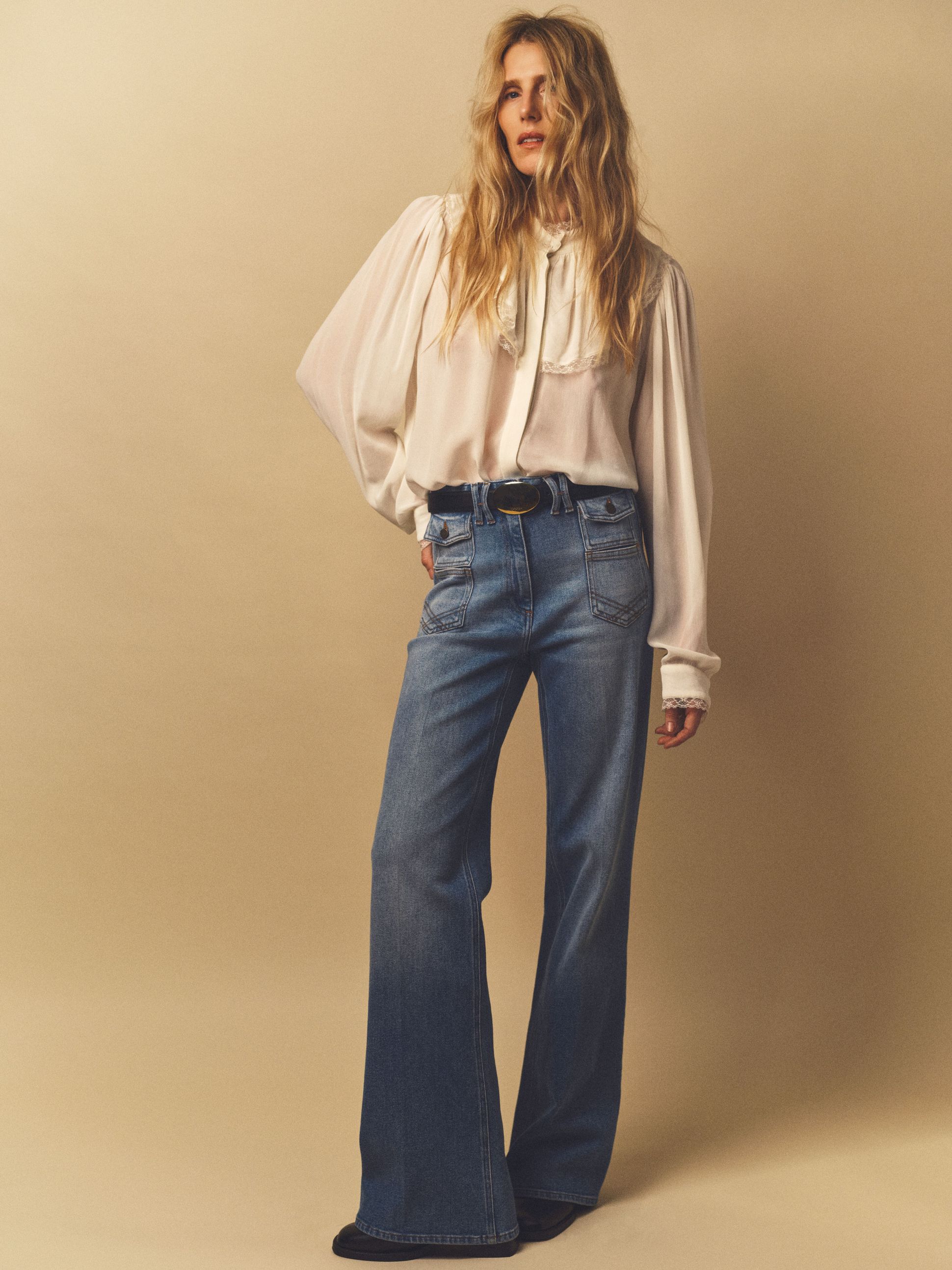 A person with long wavy hair, wearing a white blouse and high-waisted flared jeans, stands against a beige background.