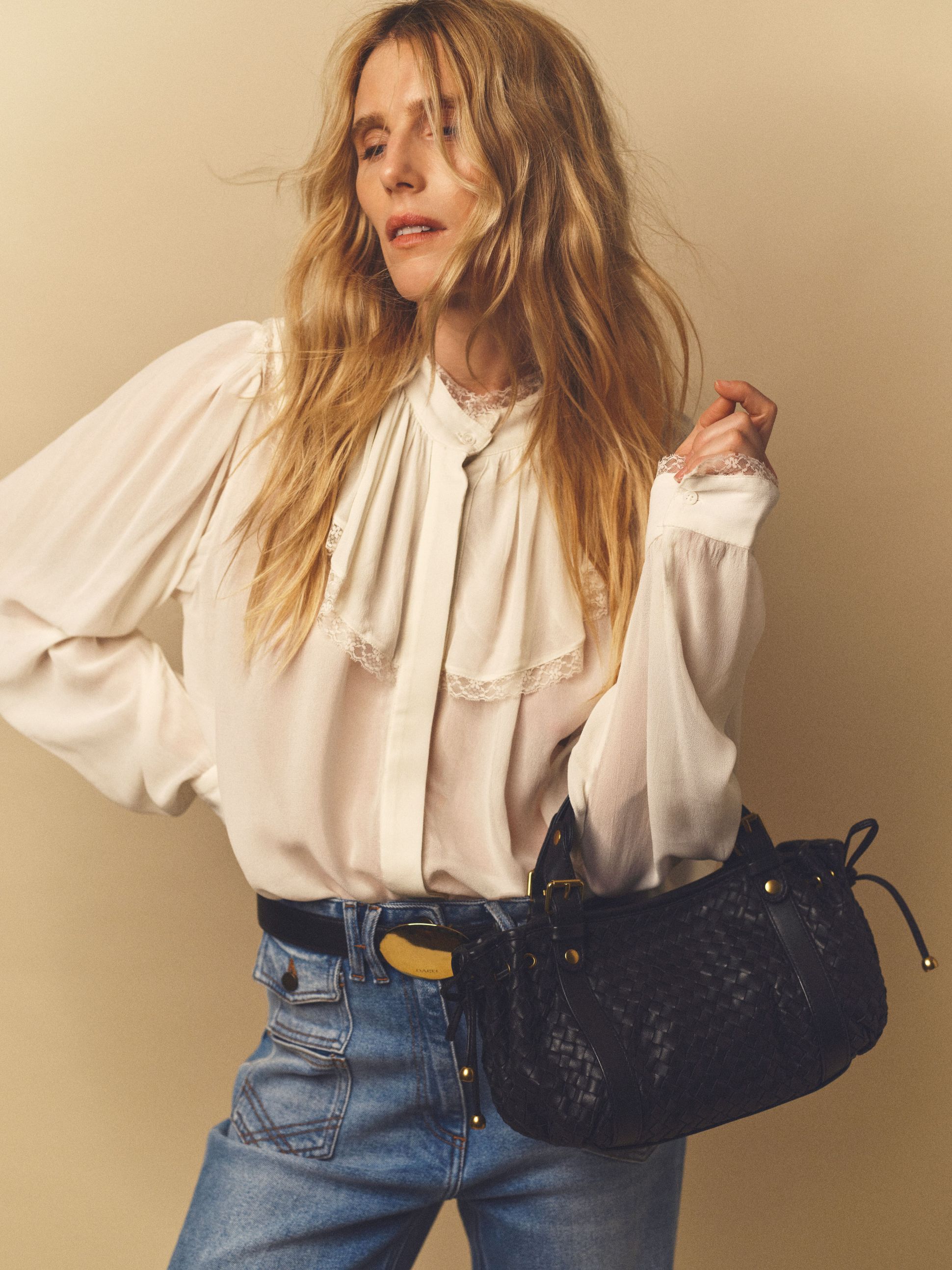 A person with long blond hair wears a white blouse and blue jeans, holding a woven black handbag, posing against a neutral background.