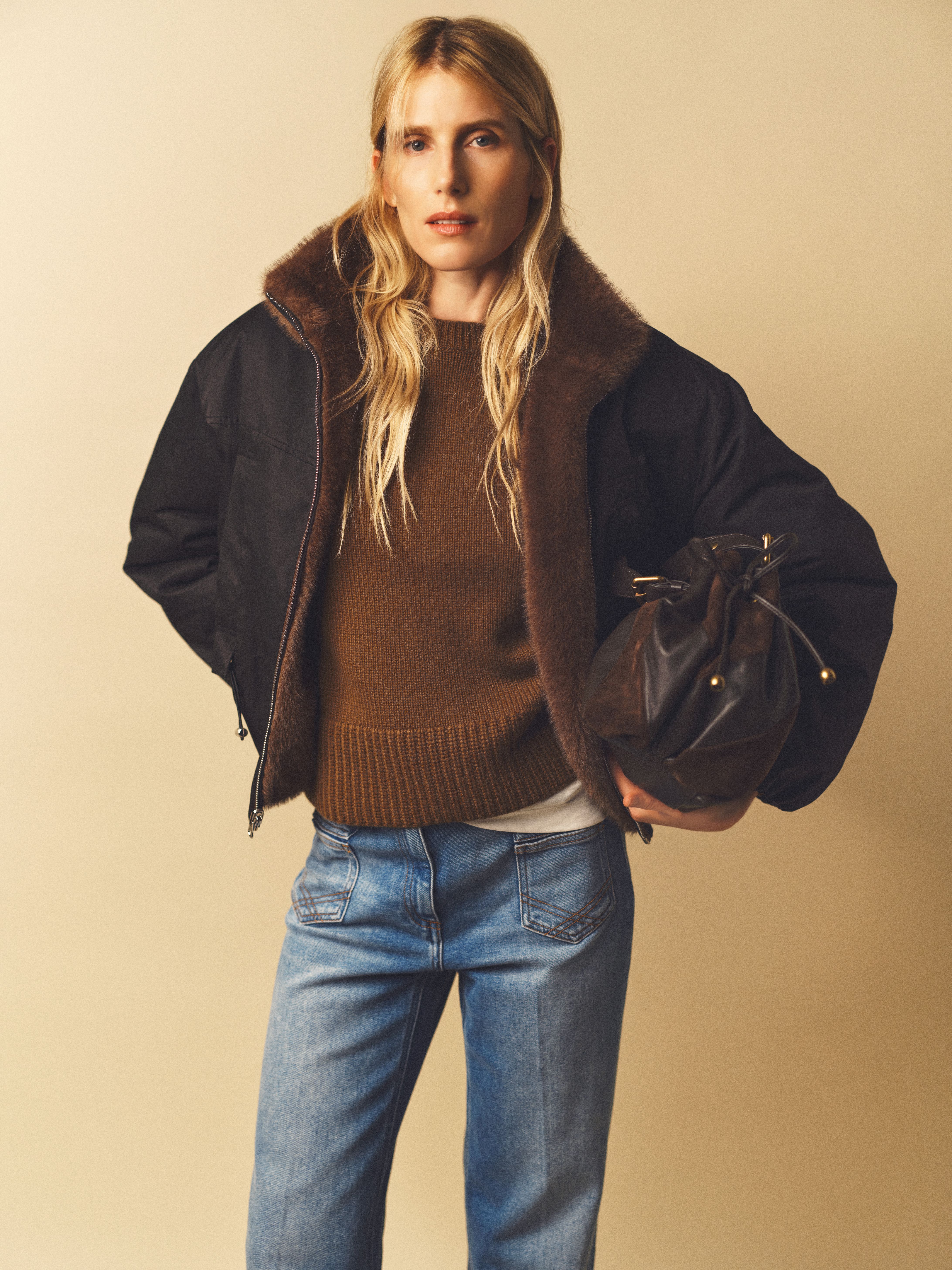 A person with long blonde hair wearing a brown sweater, dark jacket, jeans, and holding a brown bag against a beige background.