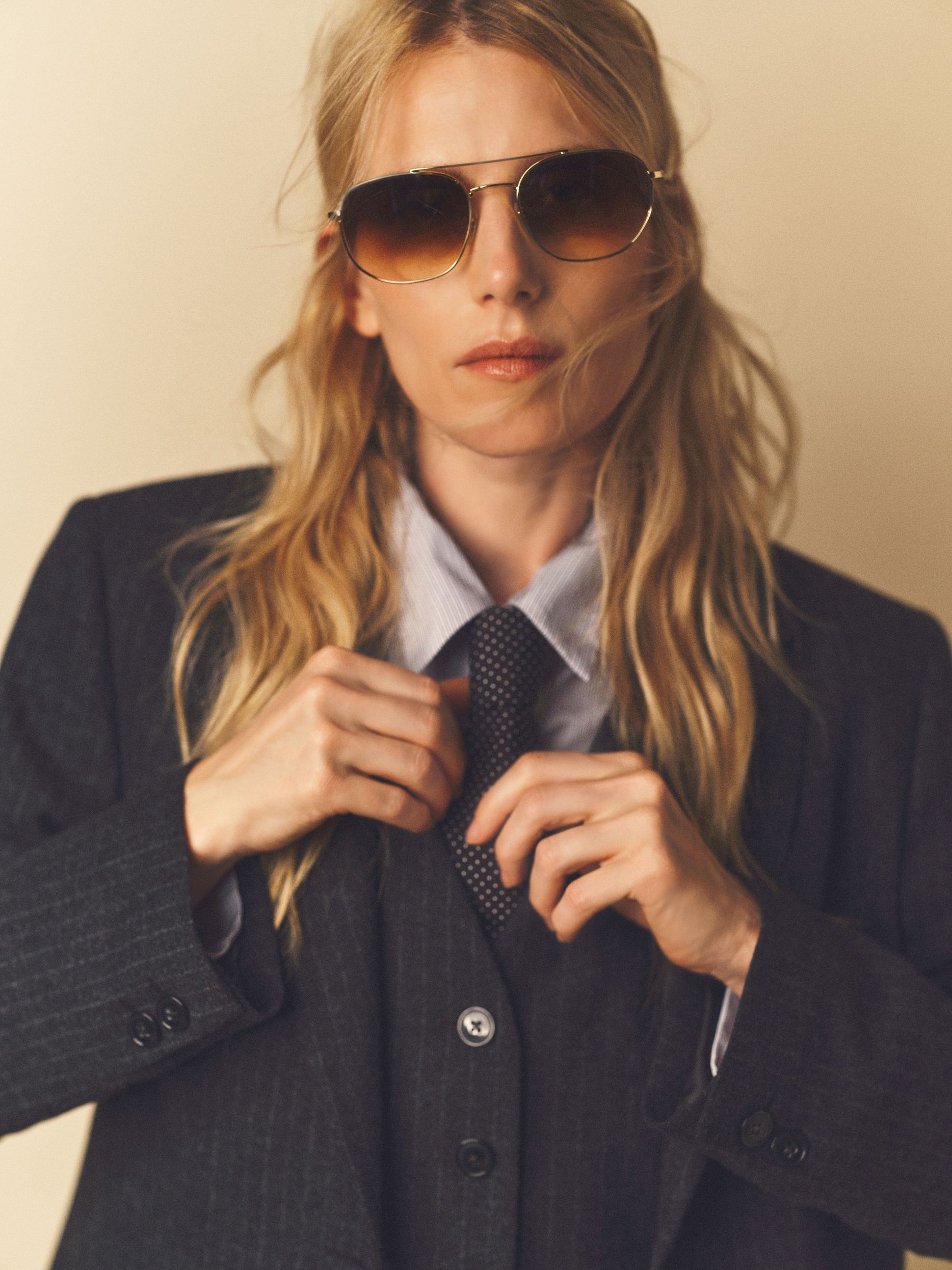 Person wearing aviator sunglasses and a dark suit adjusts their tie with a serious expression, standing against a neutral background.