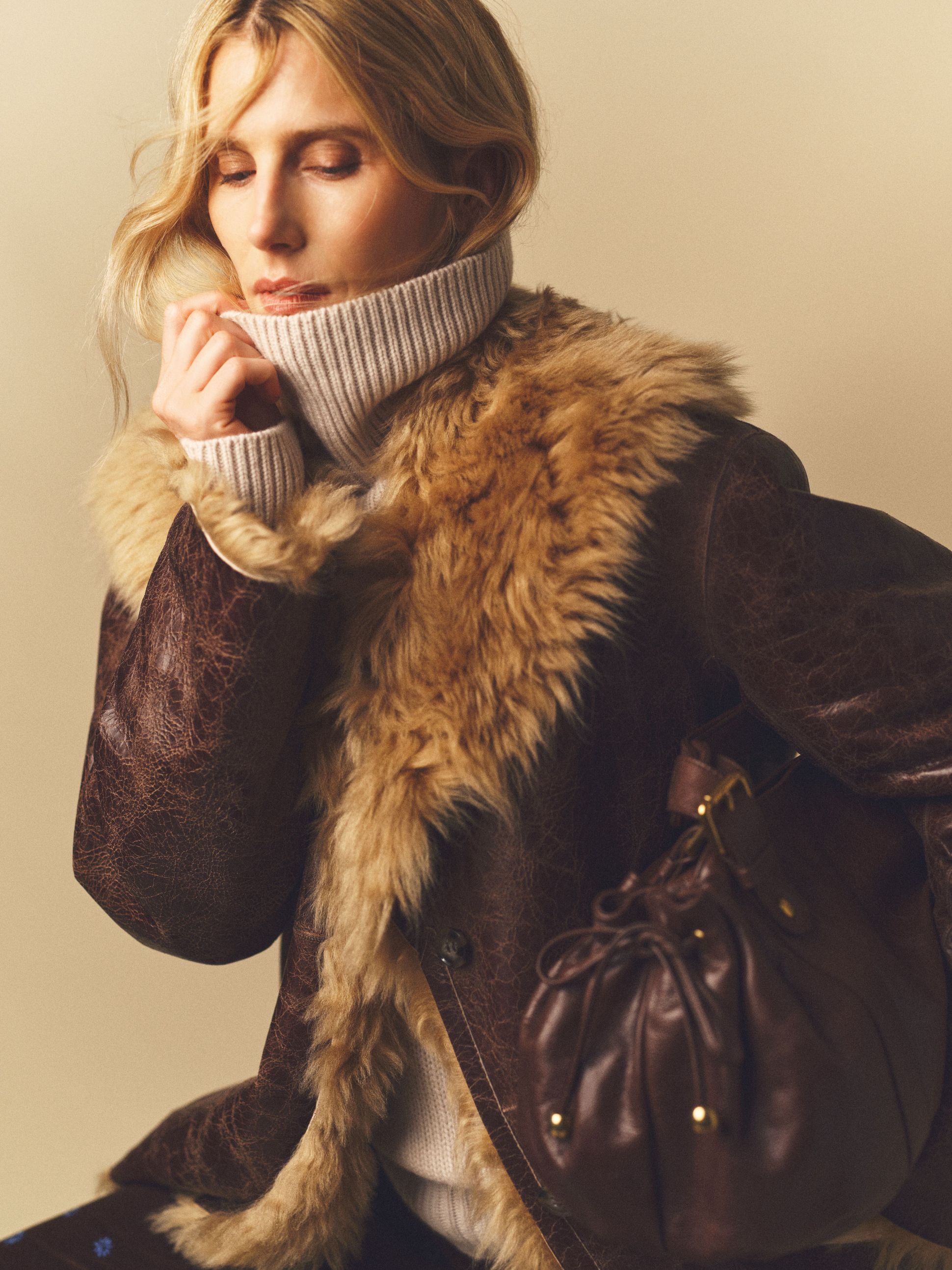 a woman with blond hair wearing a leather jacket with a fur collar and a white knit turtleneck holding a brown leather bag