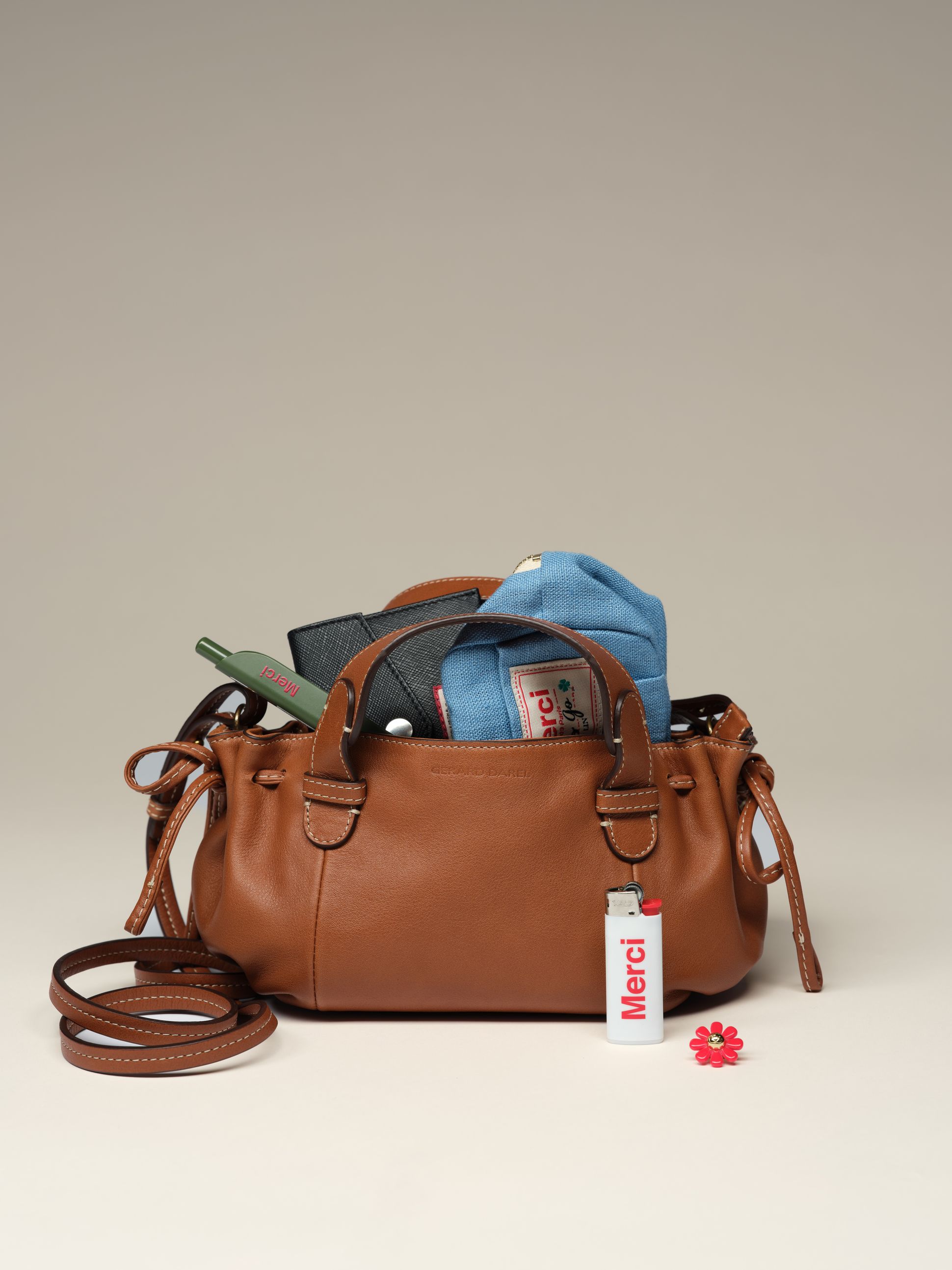 A brown leather handbag with handles, containing items like a blue fabric pouch and pen, with a white lighter and red flower accessory nearby.
