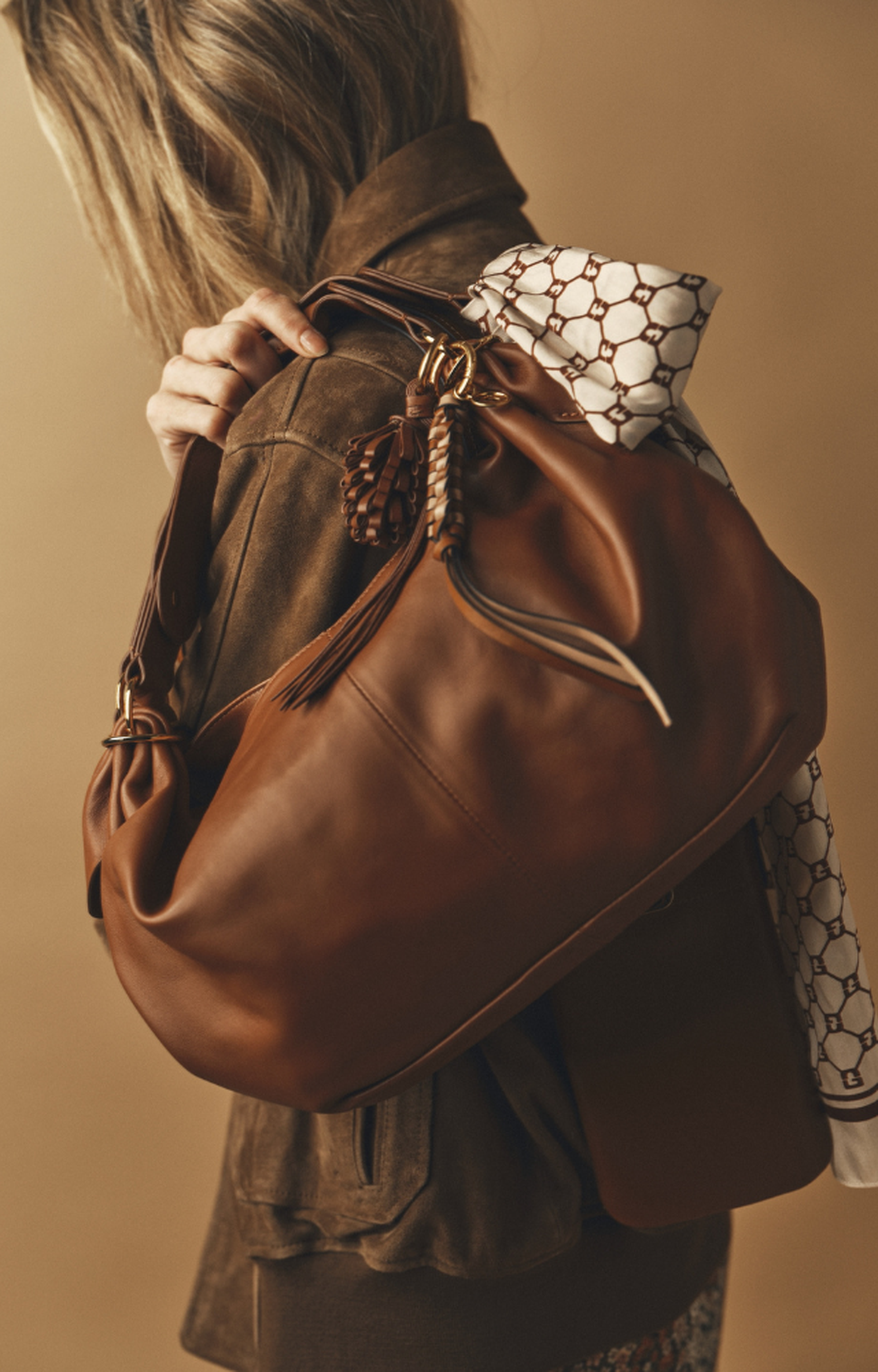 Person with shoulder-length hair holding a large brown leather bag, accessorized with a patterned scarf, wearing a brown jacket.