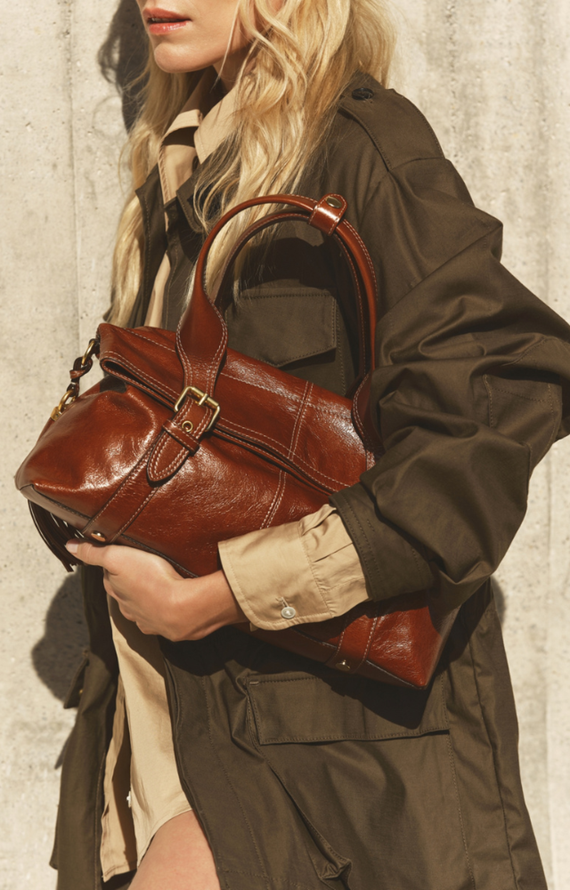A person with long blonde hair in an olive jacket holds a large brown leather bag with gold hardware against a concrete wall.