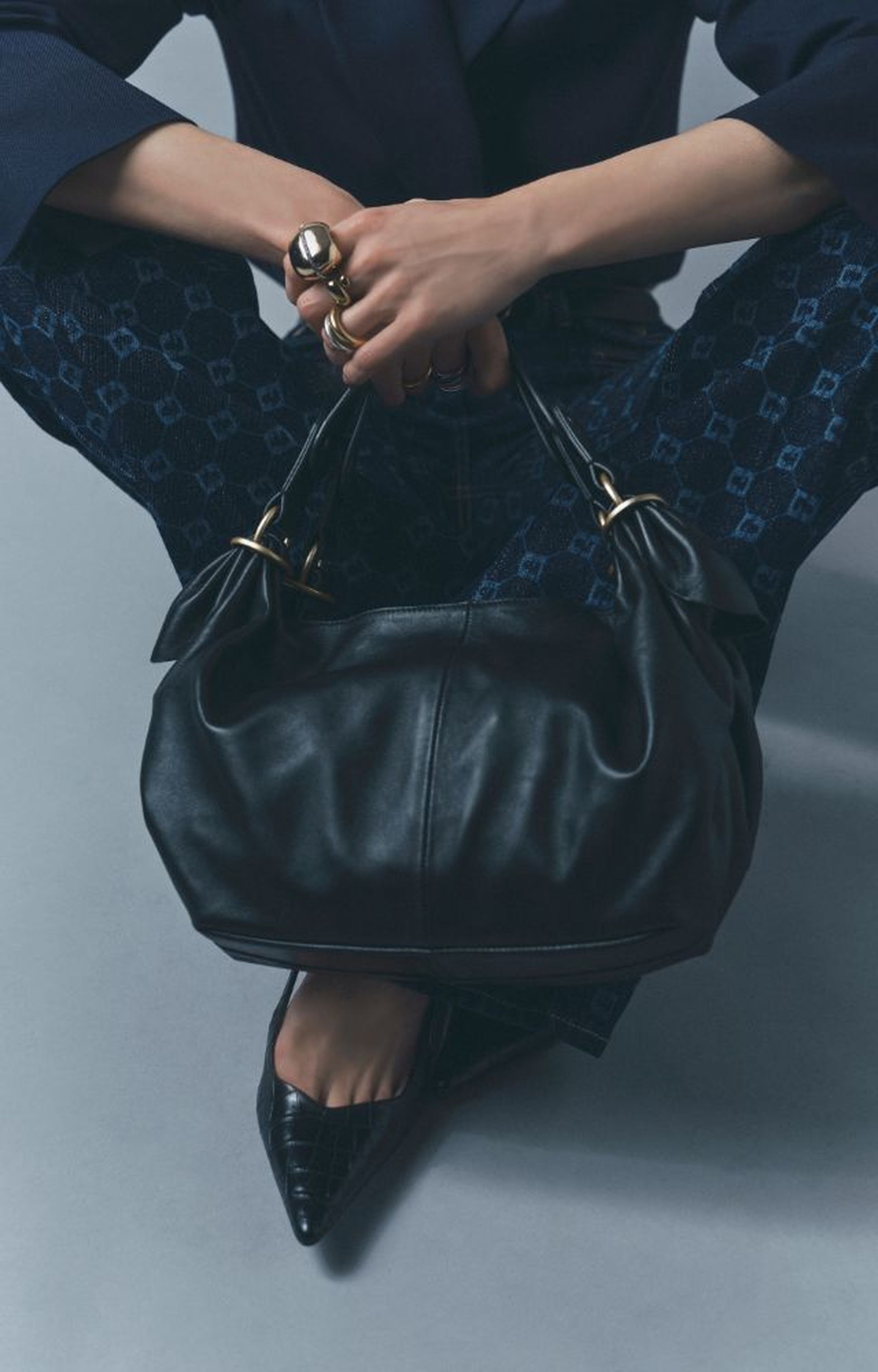 Person sitting cross-legged holding a black leather handbag, wearing blue patterned pants, a dark jacket, and pointy black shoes.