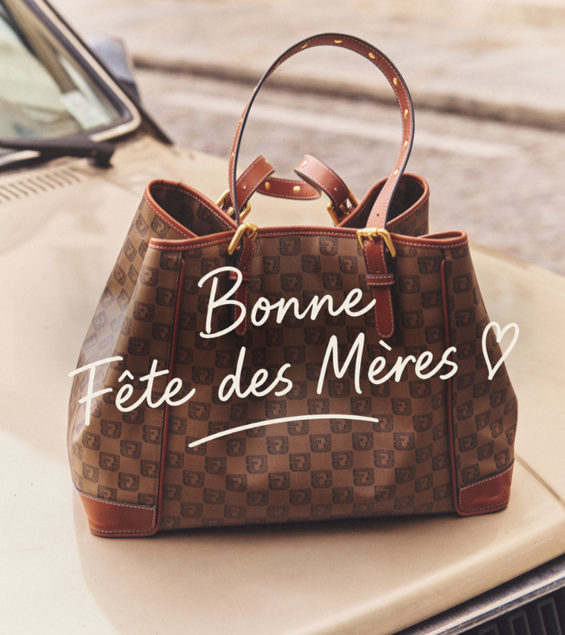 A patterned brown handbag with leather accents sits on a car hood. Text reads "Bonne Fête des Mères" with a heart symbol.