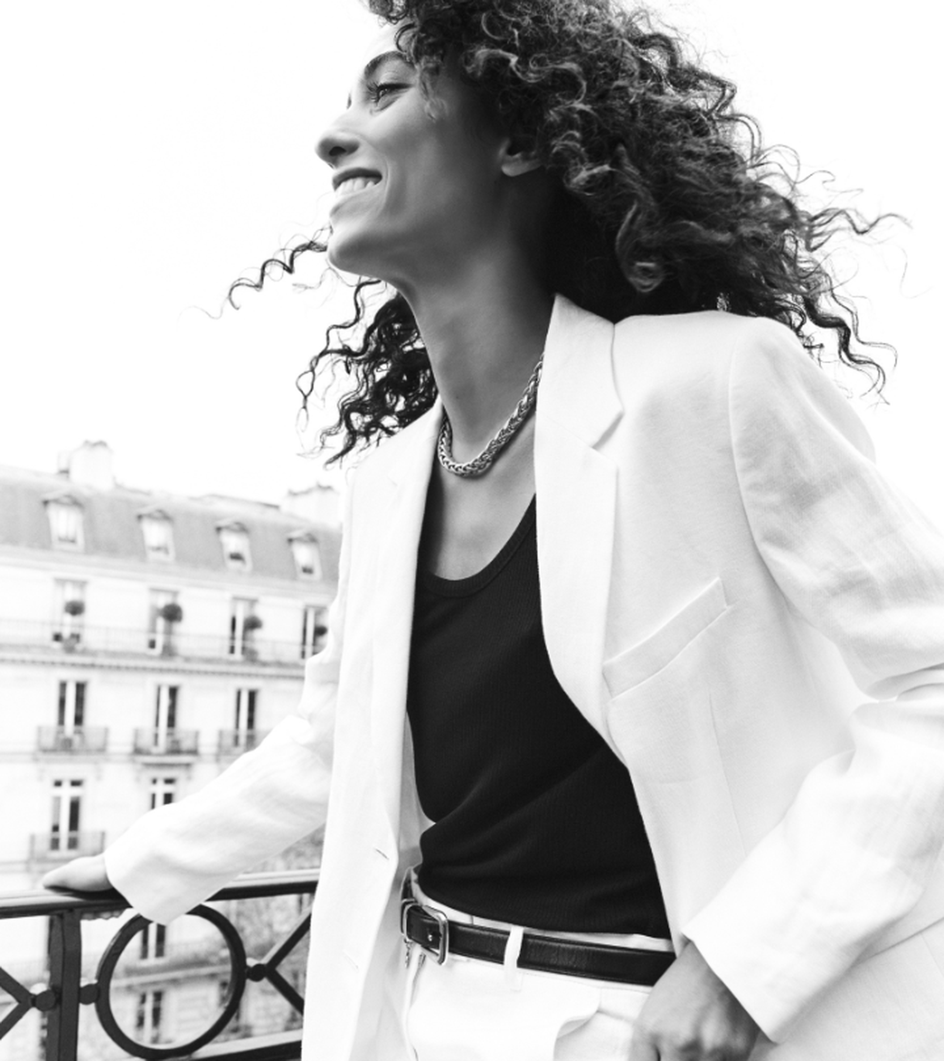 Smiling person in a white blazer and black top, standing on a balcony with a view of classic buildings in the background. Black and white image.