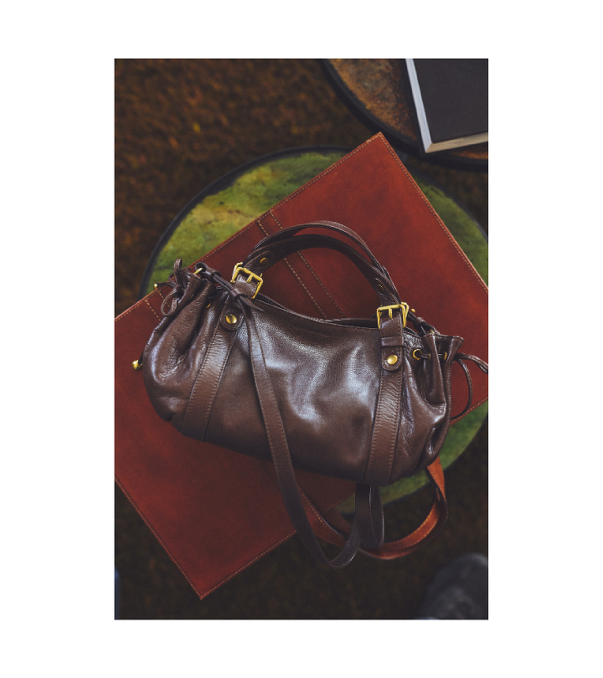 A brown leather handbag with gold hardware sits on a red folder on a round table, with a closed book nearby.