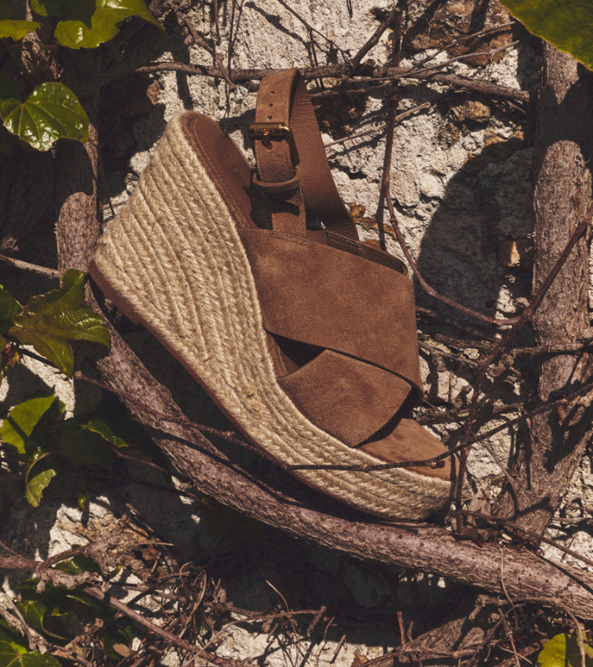 Brown suede wedge sandal with jute platform, strapped around a weathered wall with ivy and branches.