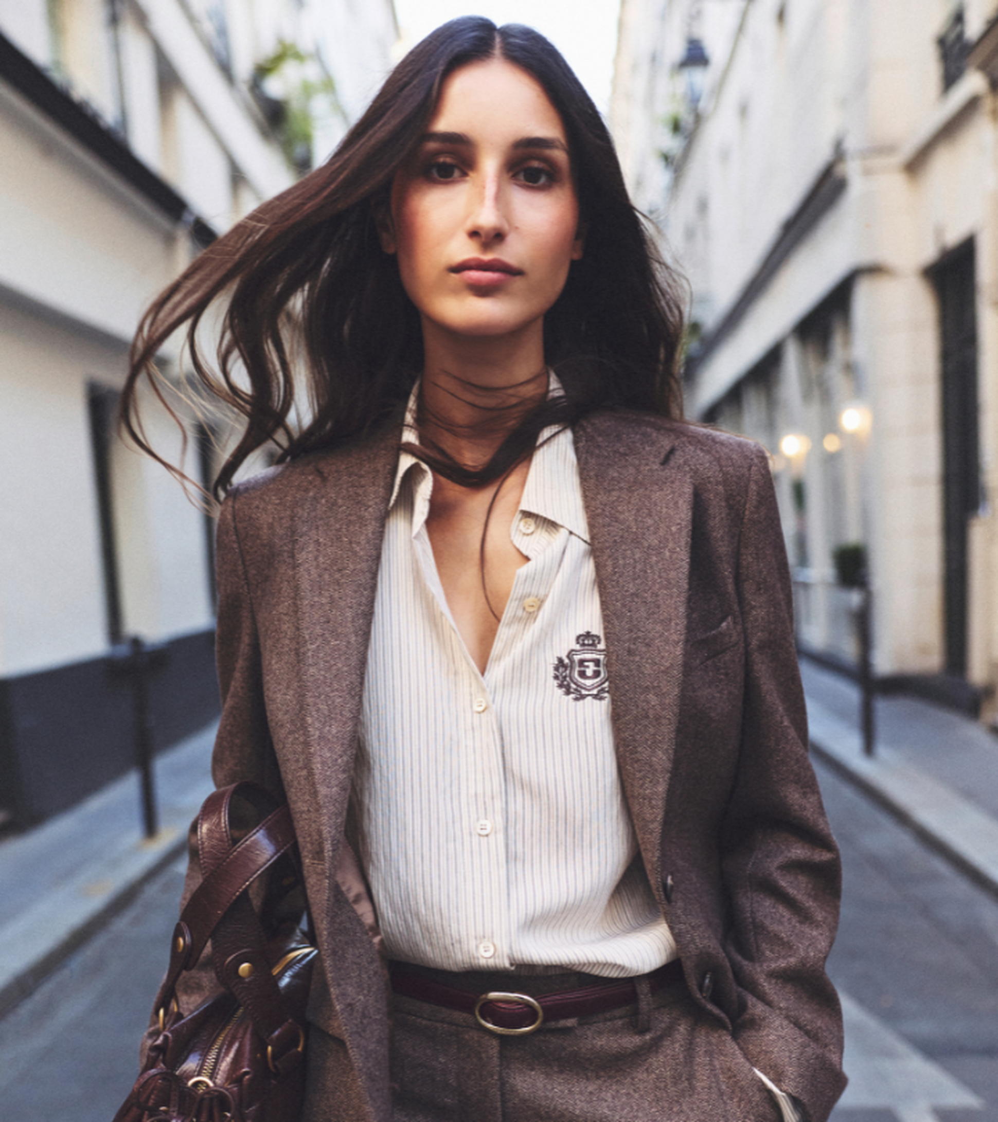 A woman in a stylish brown suit and striped shirt walking down a city street, with long hair flowing in the wind.