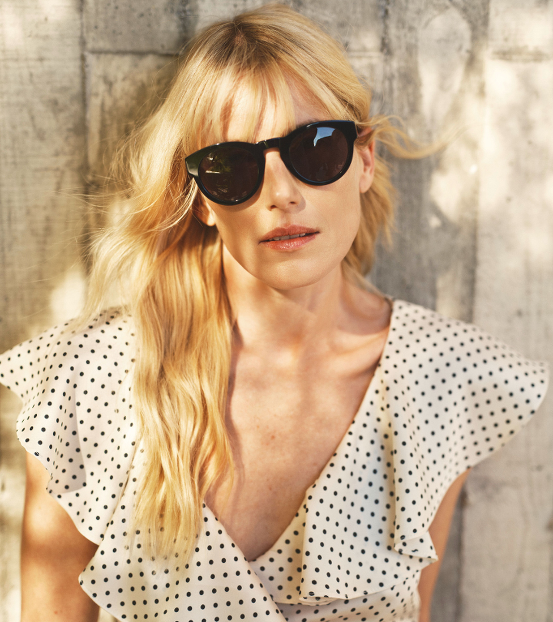 Woman with long blonde hair and sunglasses in polka dot dress, standing against a concrete wall.