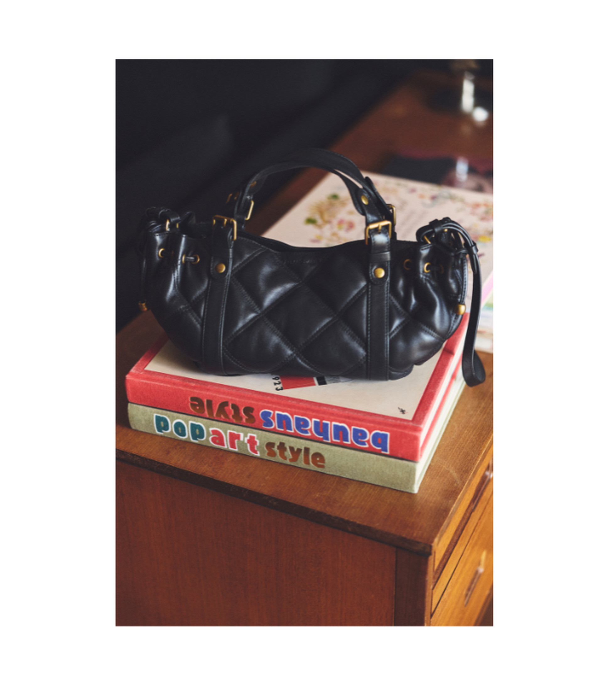 Black quilted handbag on a stack of colorful pop art style books on a wooden table.