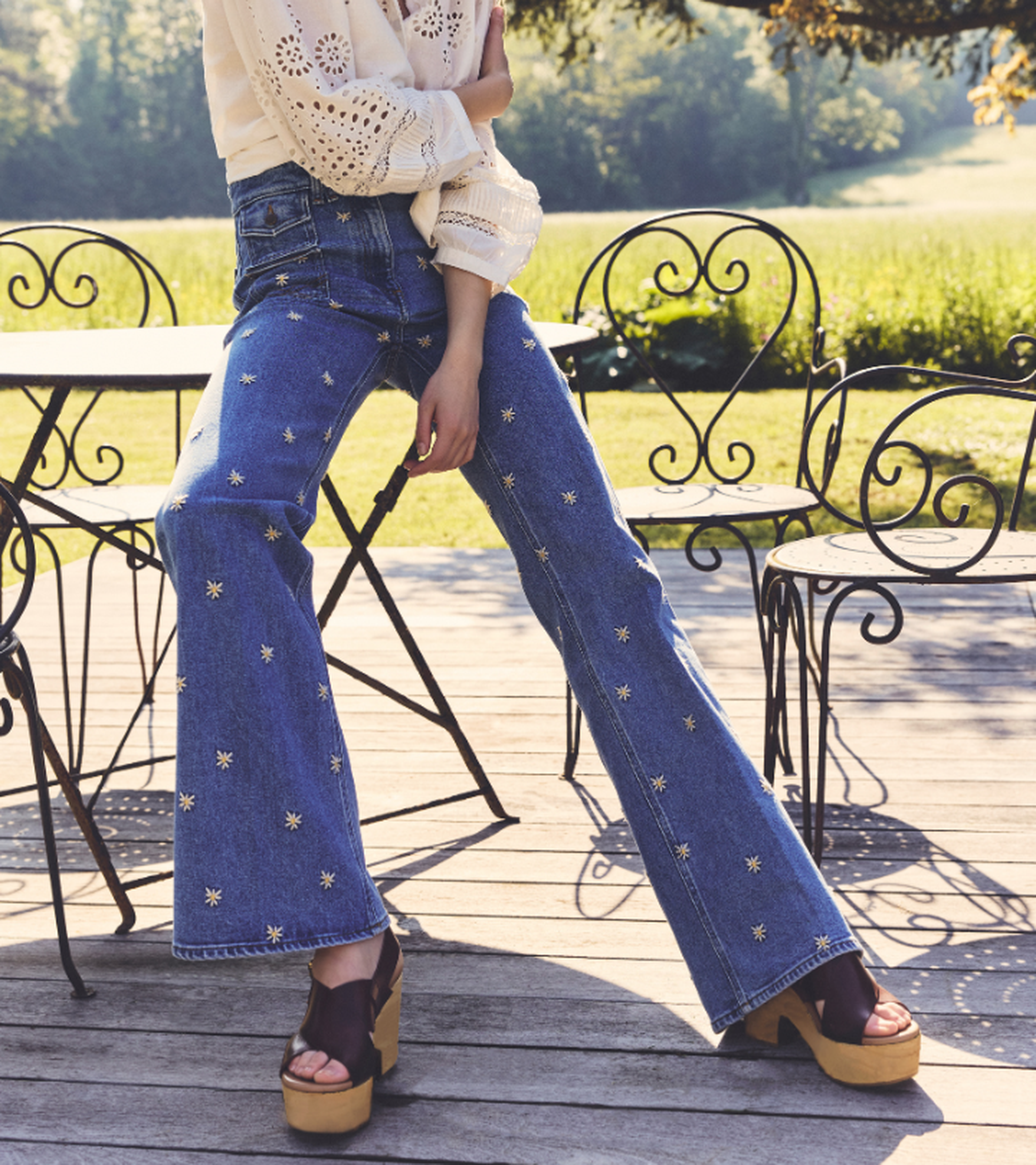 Person in embroidered jeans and platform sandals sits casually on an outdoor patio with iron furniture, set against a lush green landscape.