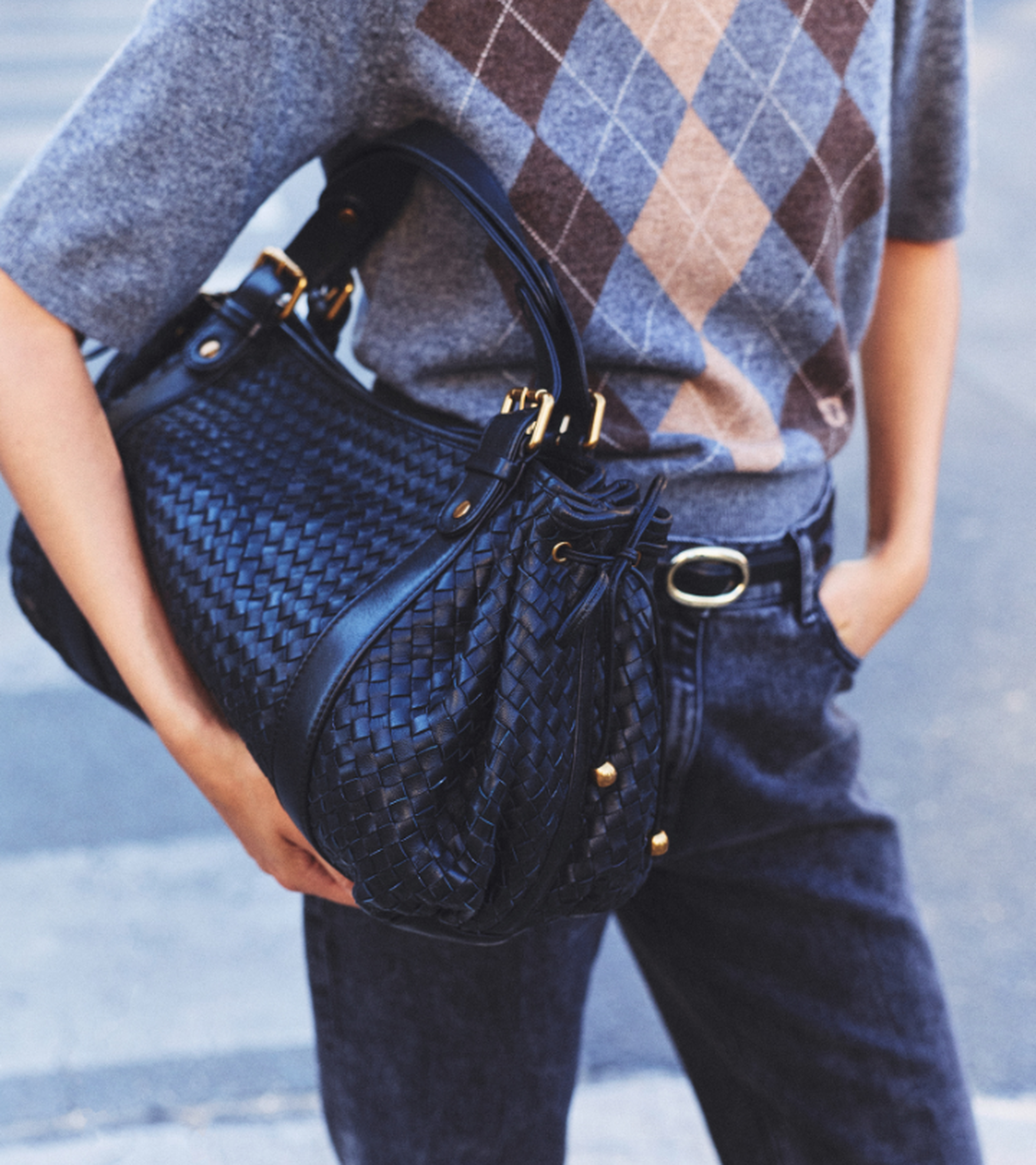 Person wearing an argyle sweater and jeans holds a large, woven black handbag on their arm while standing on a sidewalk.