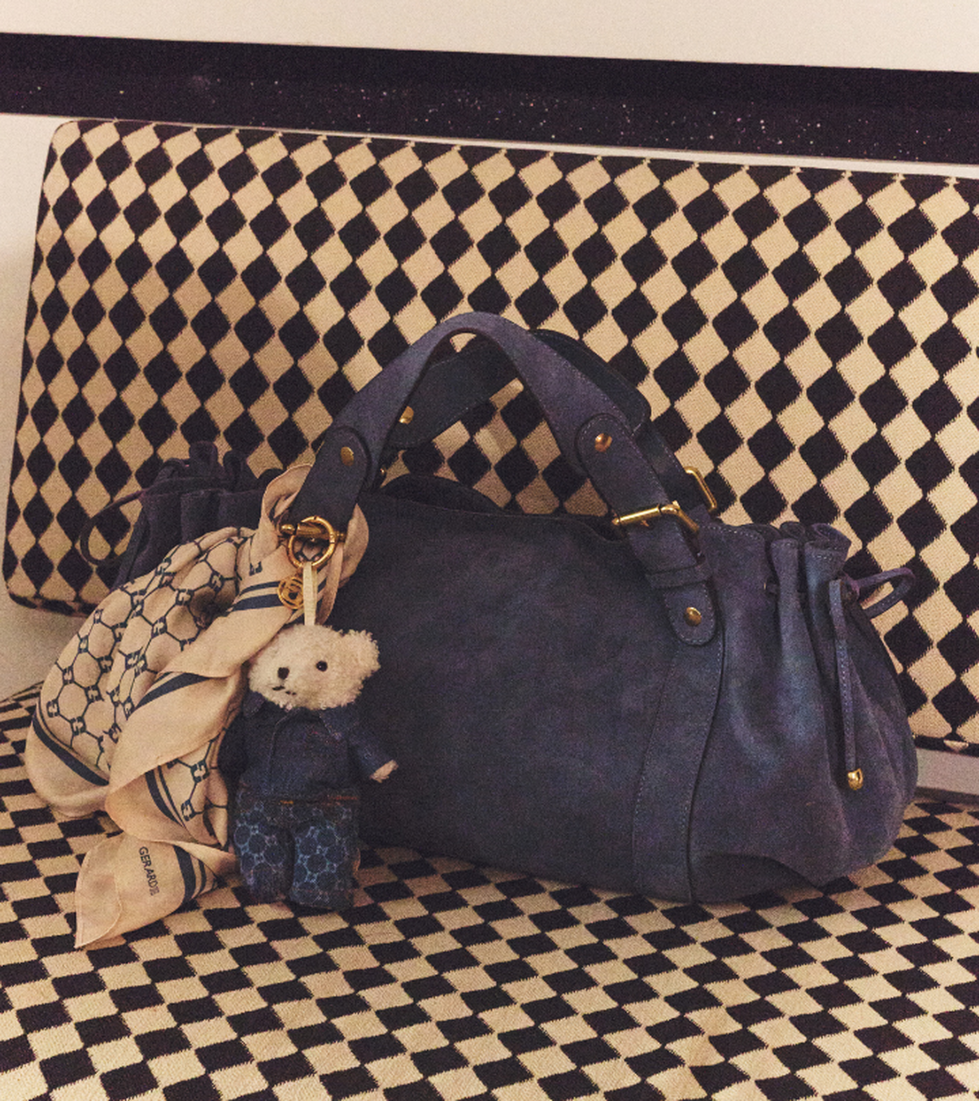 Blue handbag with a teddy bear charm and scarf, placed on a black and white checkered bench.