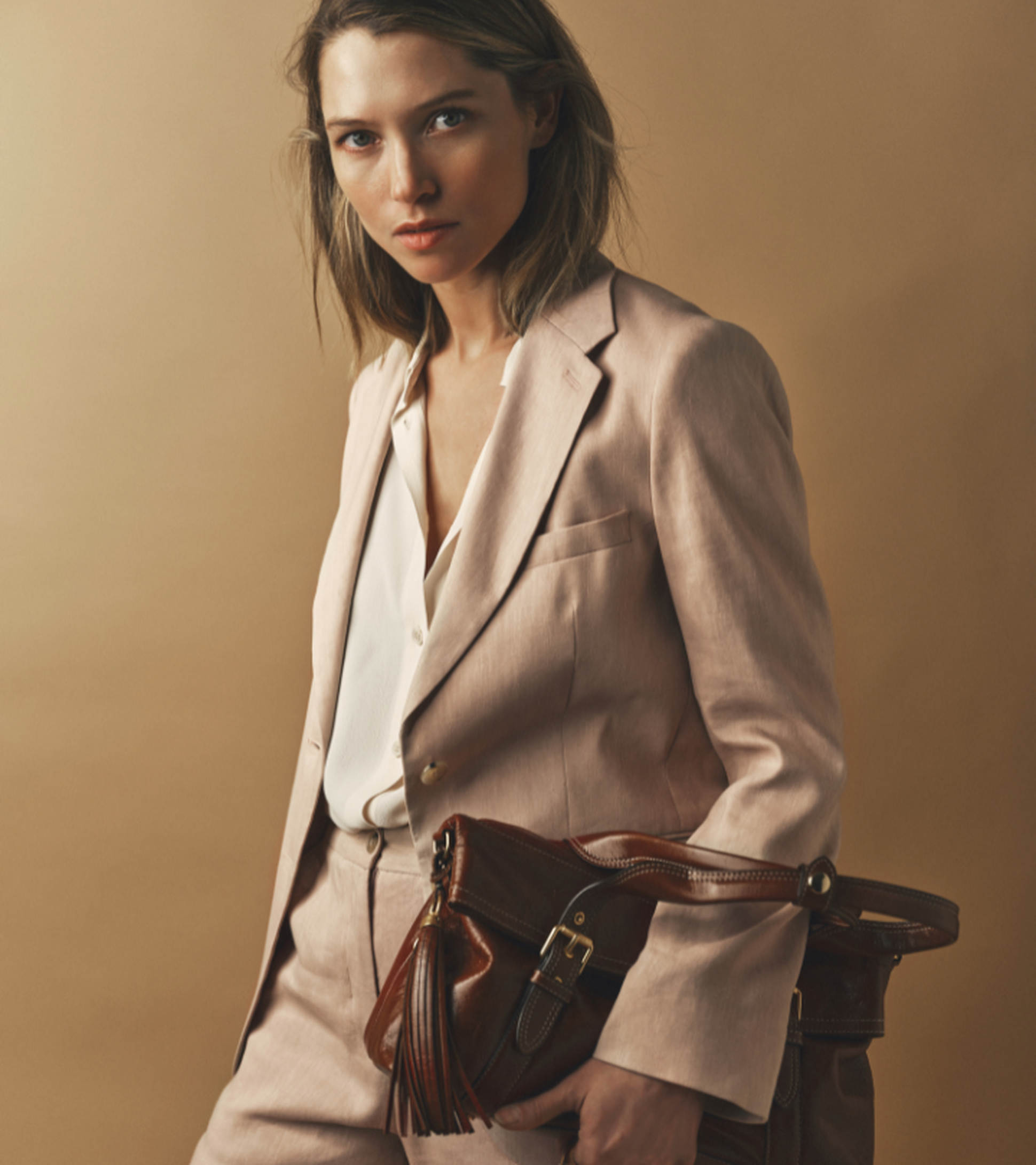Woman in a pink suit holding a brown leather bag, standing against a beige background, looking at the camera.