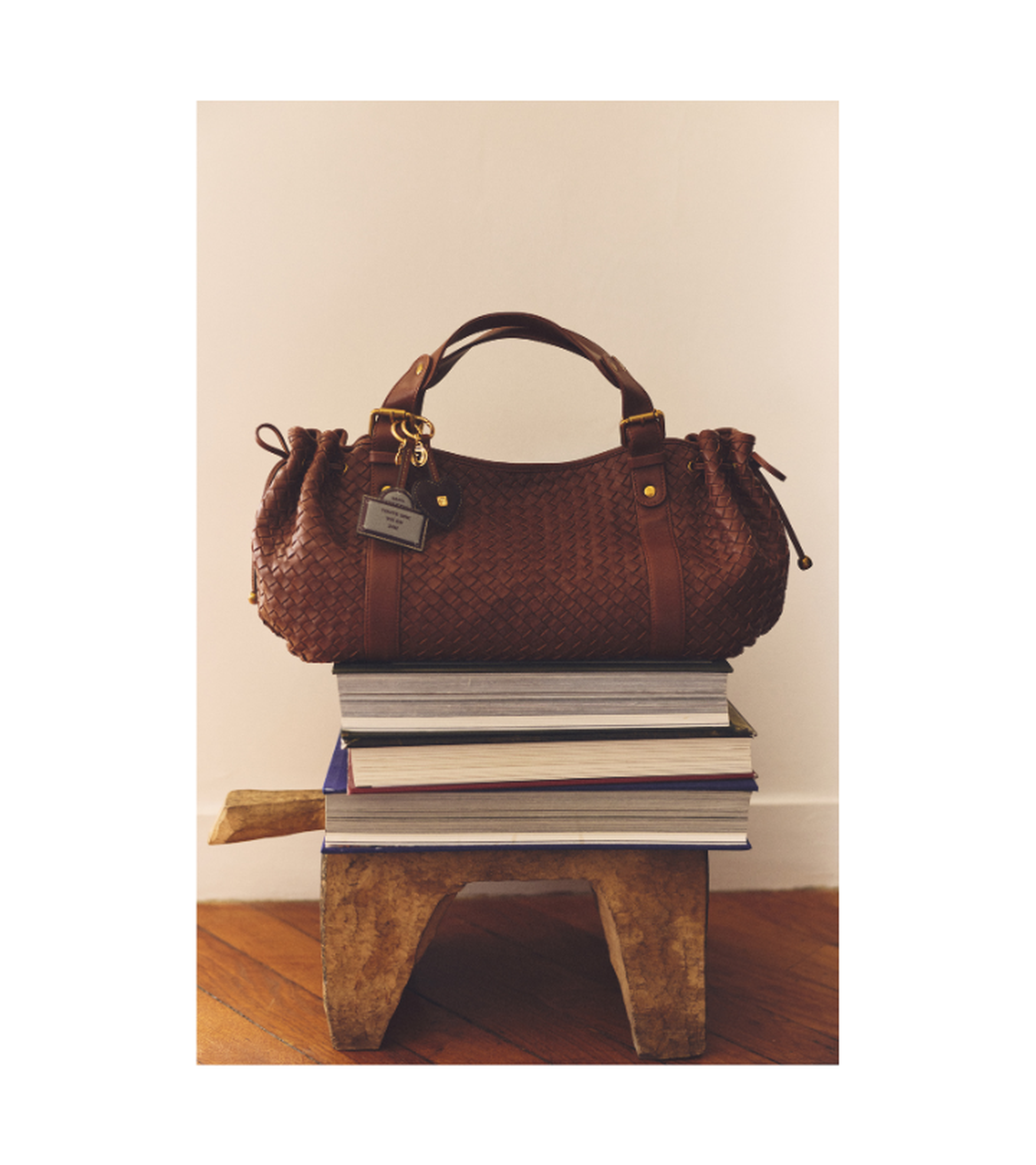 A woven brown handbag sits on top of a stack of books placed on a small wooden stool against a plain background.