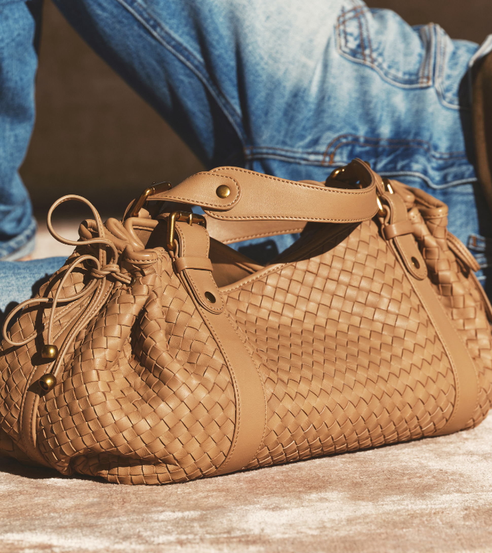 A tan leather woven handbag with drawstring details, set against a background of light denim and concrete.