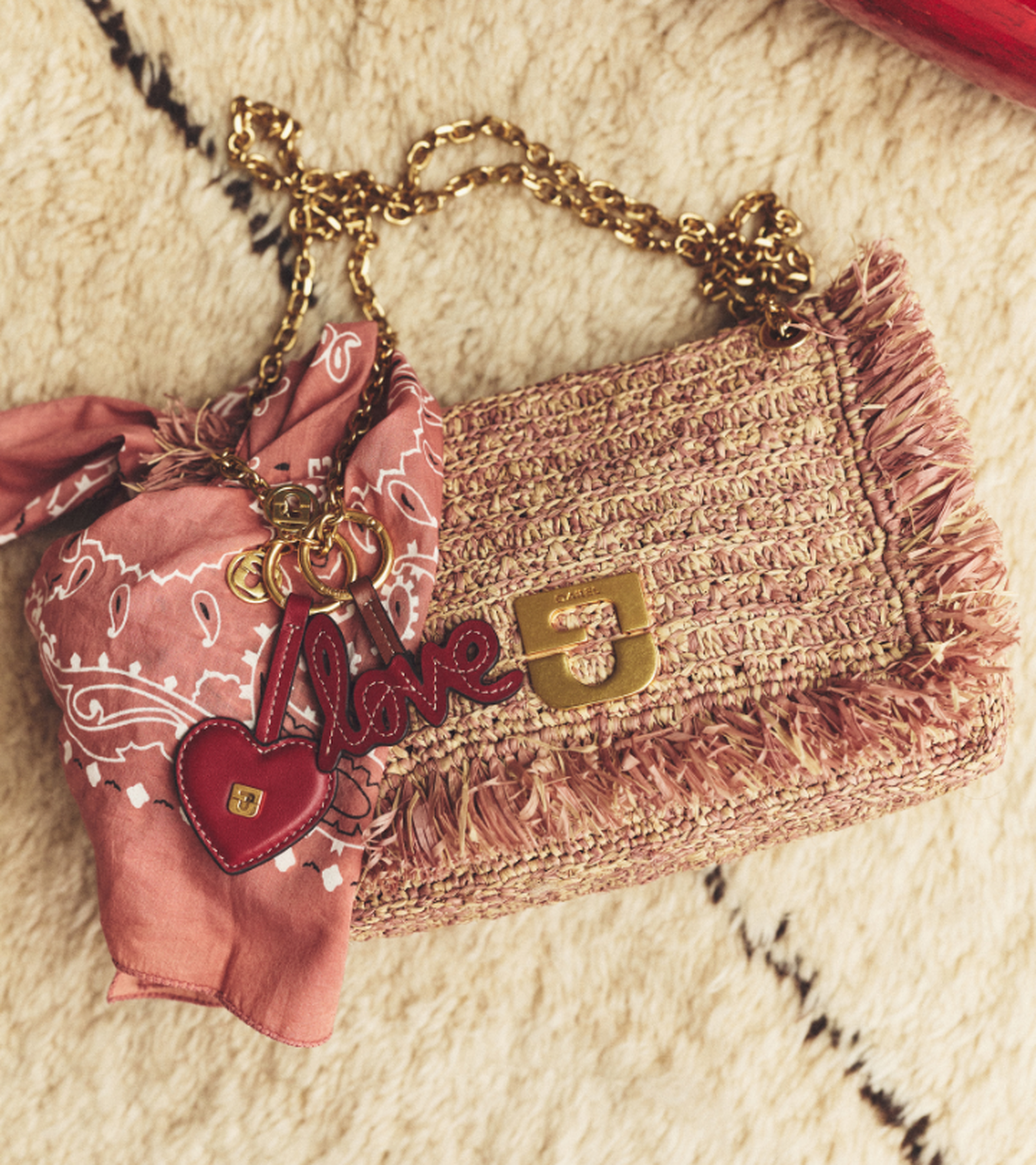 A woven purse with a gold chain, pink bandana, and "love" charm lies on a textured surface.