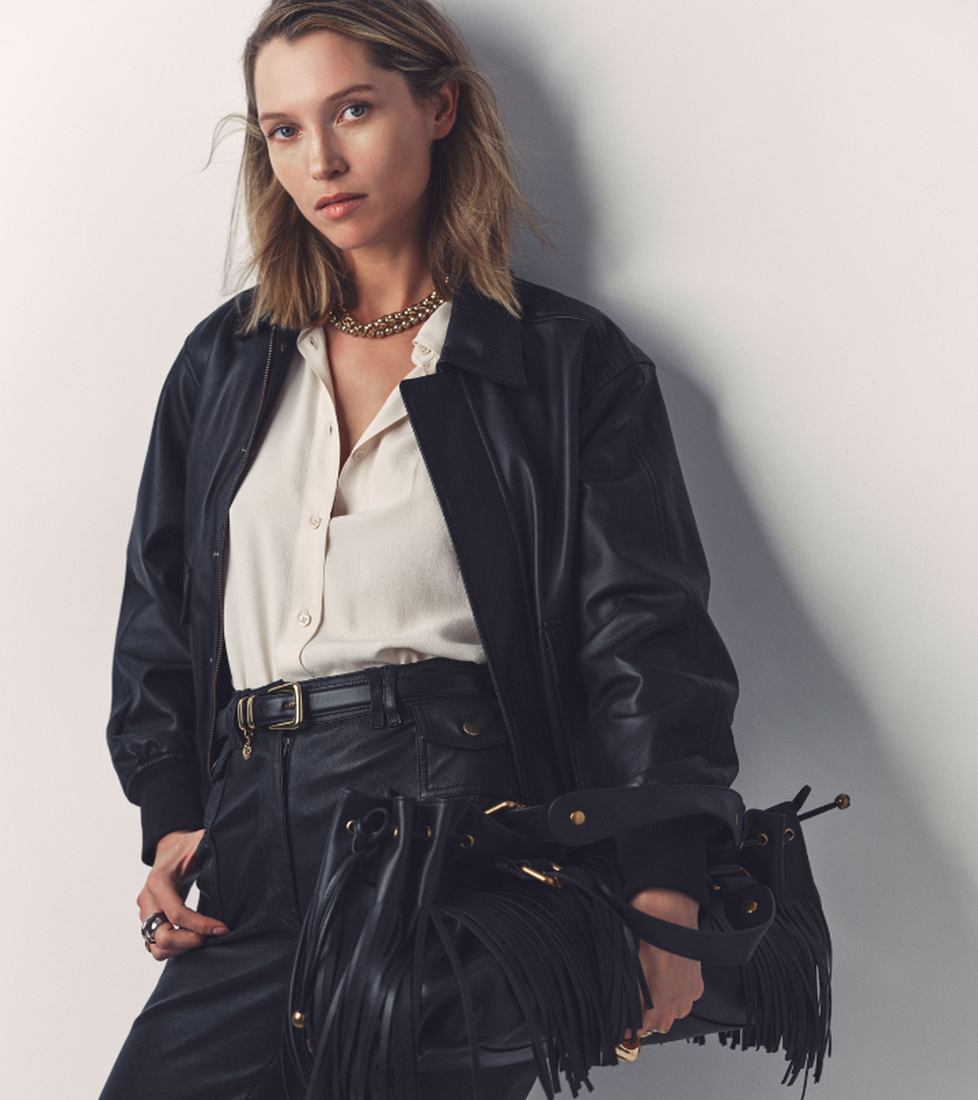Woman in black leather jacket and pants holding a fringed black handbag. She wears a cream blouse and gold necklace, standing against a plain backdrop.