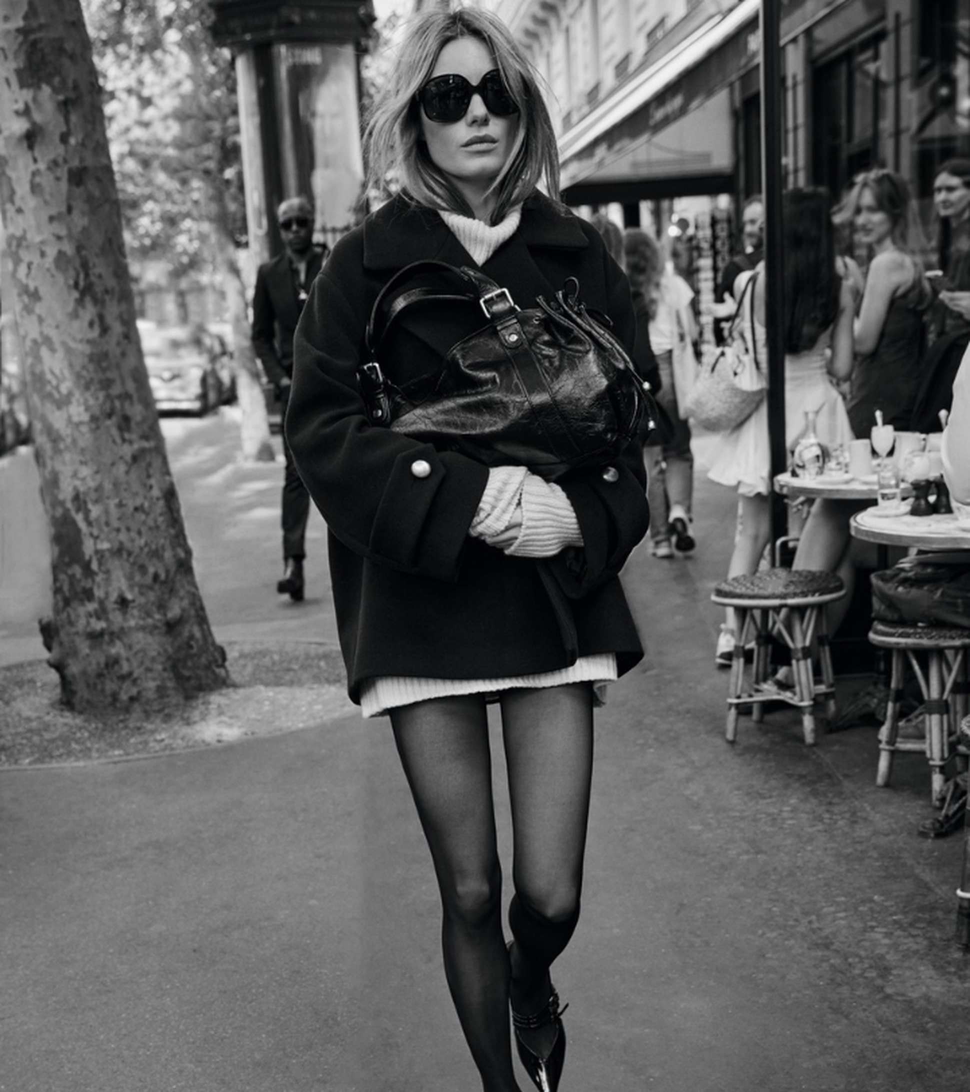A woman in sunglasses and a black coat walks on a city street carrying a handbag, with outdoor cafe tables and pedestrians in the background.