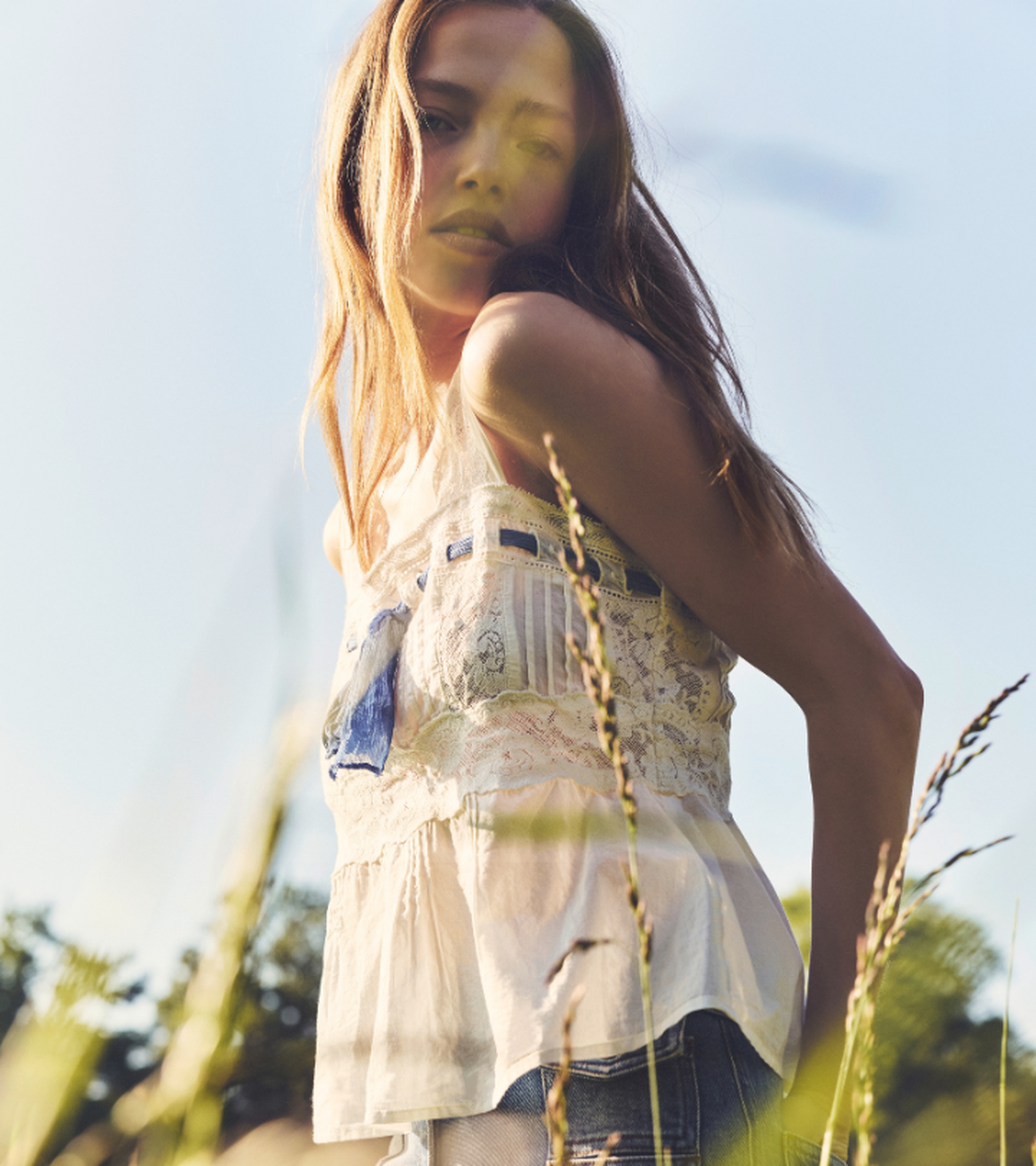 Young woman in a sleeveless top and jean shorts stands in a sunlit field with tall grass, looking towards the camera.