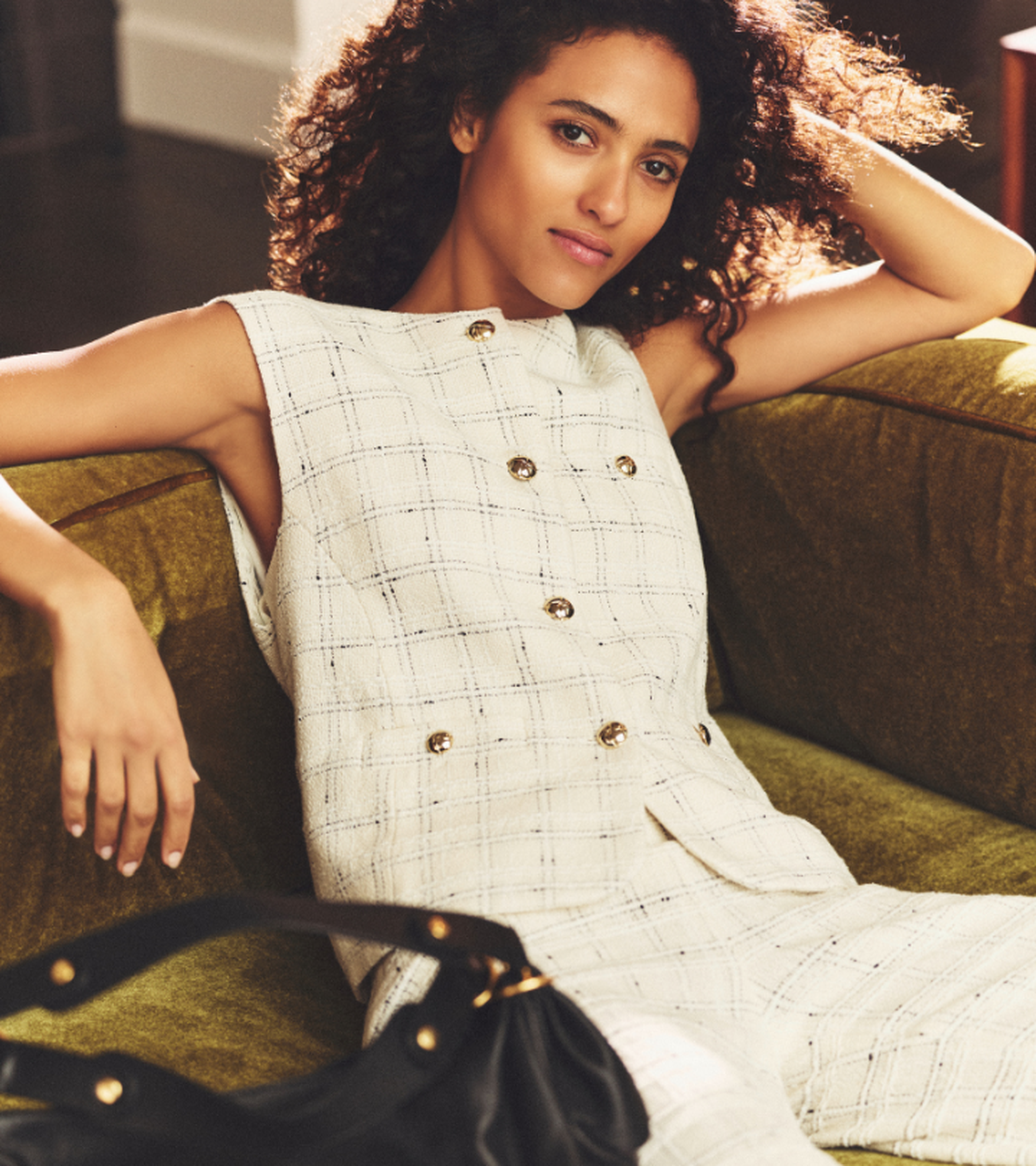 Woman with curly hair in a white plaid outfit, lounging on a green couch with a black handbag in the foreground.