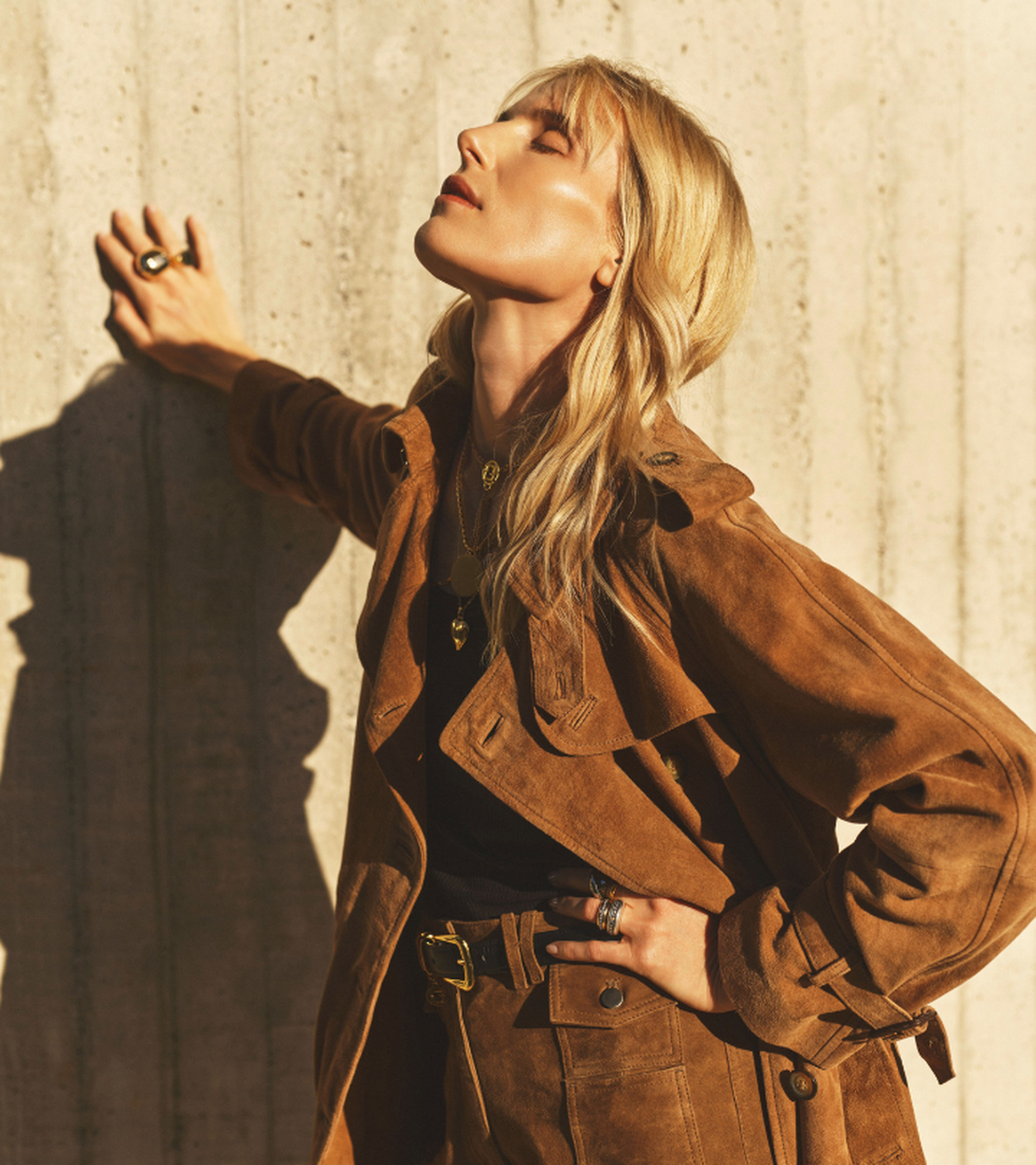 Woman with blonde hair leans against a sunlit wall, wearing a brown suede jacket and black top, eyes closed in a relaxed pose.
