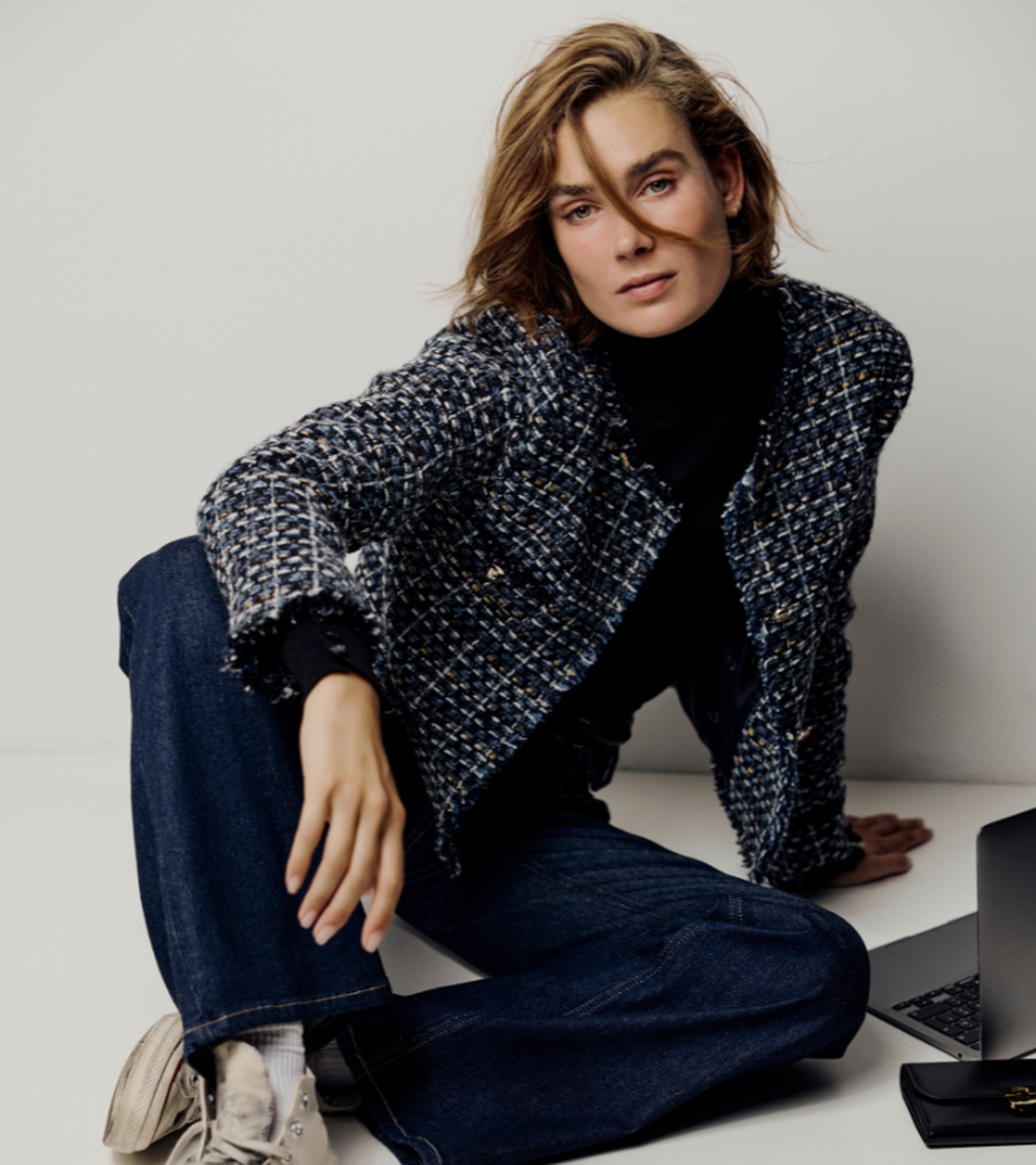 Person sitting on the floor, wearing a patterned jacket and jeans, with a neutral expression. A laptop is partially visible nearby.