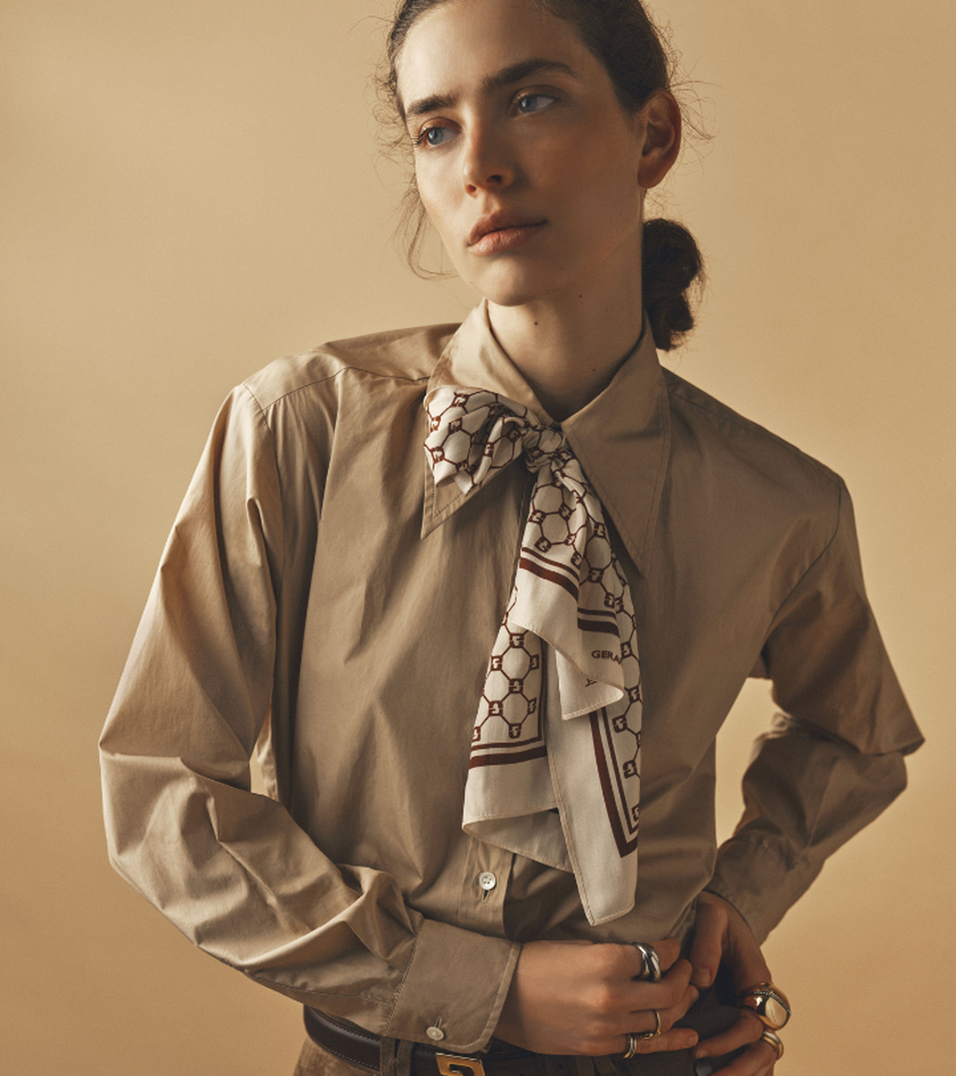 Person in a beige shirt with a patterned scarf tied at the collar, looking to the side, wearing multiple rings, on a neutral background.
