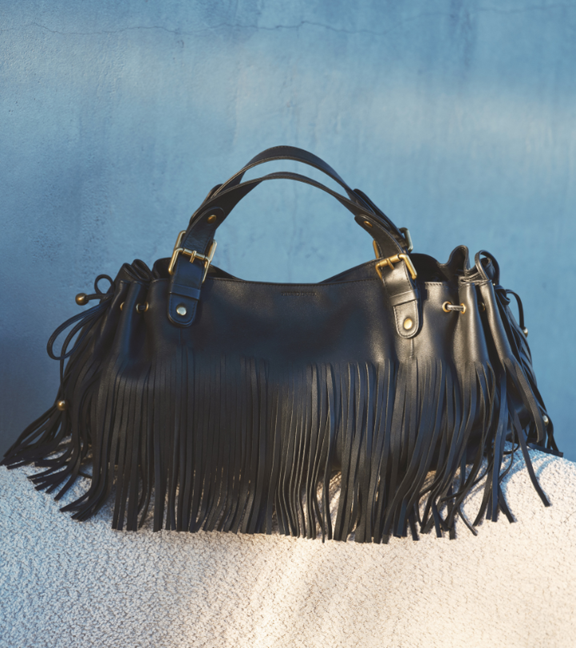 Black leather handbag with fringe details, gold hardware, and a double handle, resting against a textured surface with blue background.