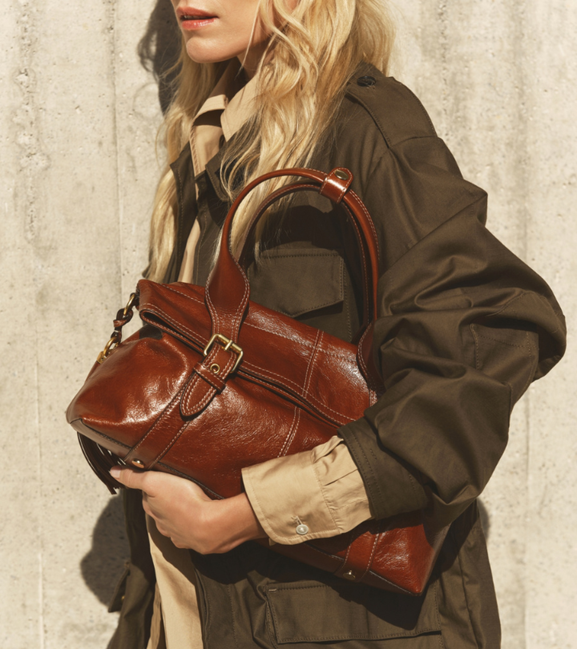 Woman in a brown coat holds a leather handbag under arm against a textured concrete wall.