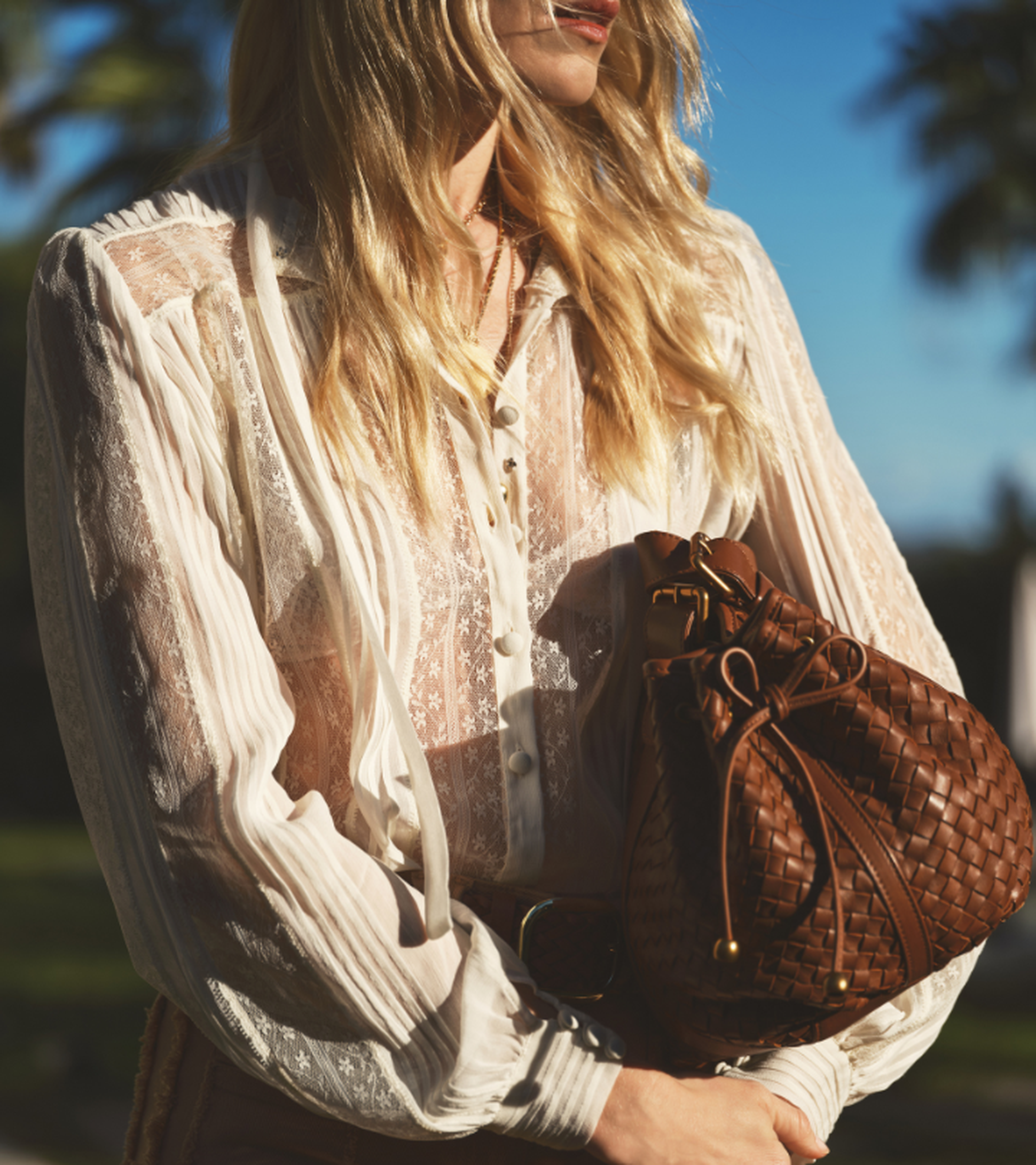 A woman in a sheer white lace blouse holds a brown woven leather bag, standing outdoors with blurred greenery and blue sky in the background.