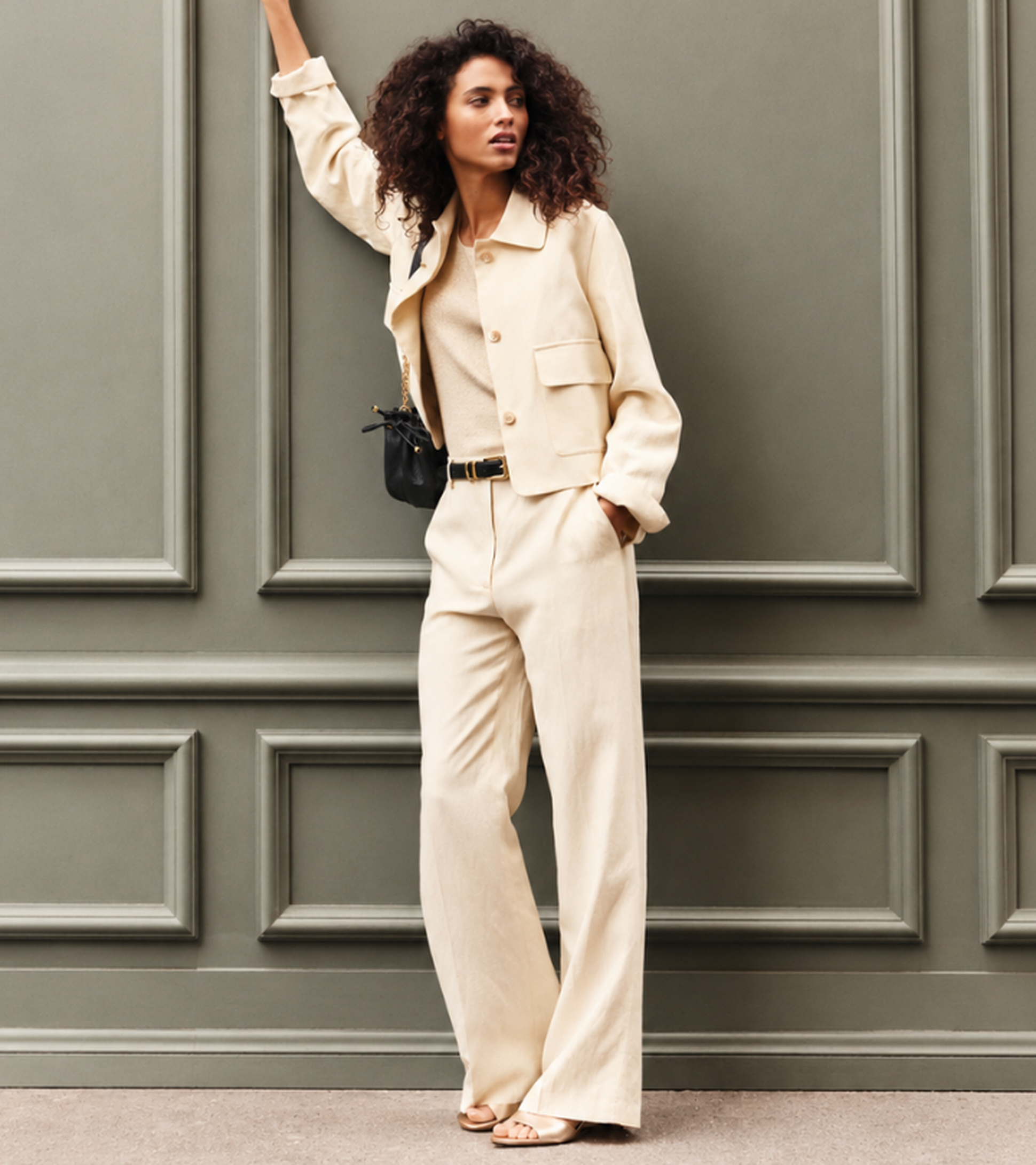 Woman in a beige outfit with a jacket and wide-leg pants leans casually against a decorative olive-green wall, holding a small black purse.