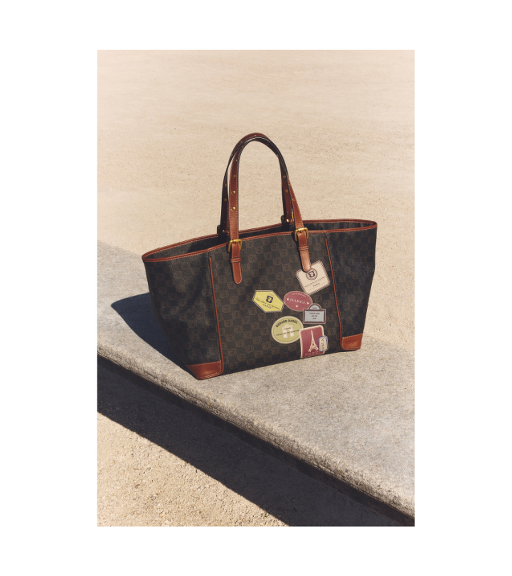 A patterned tote bag with leather handles is adorned with colorful travel-themed patches, sitting on a concrete ledge under the sun.