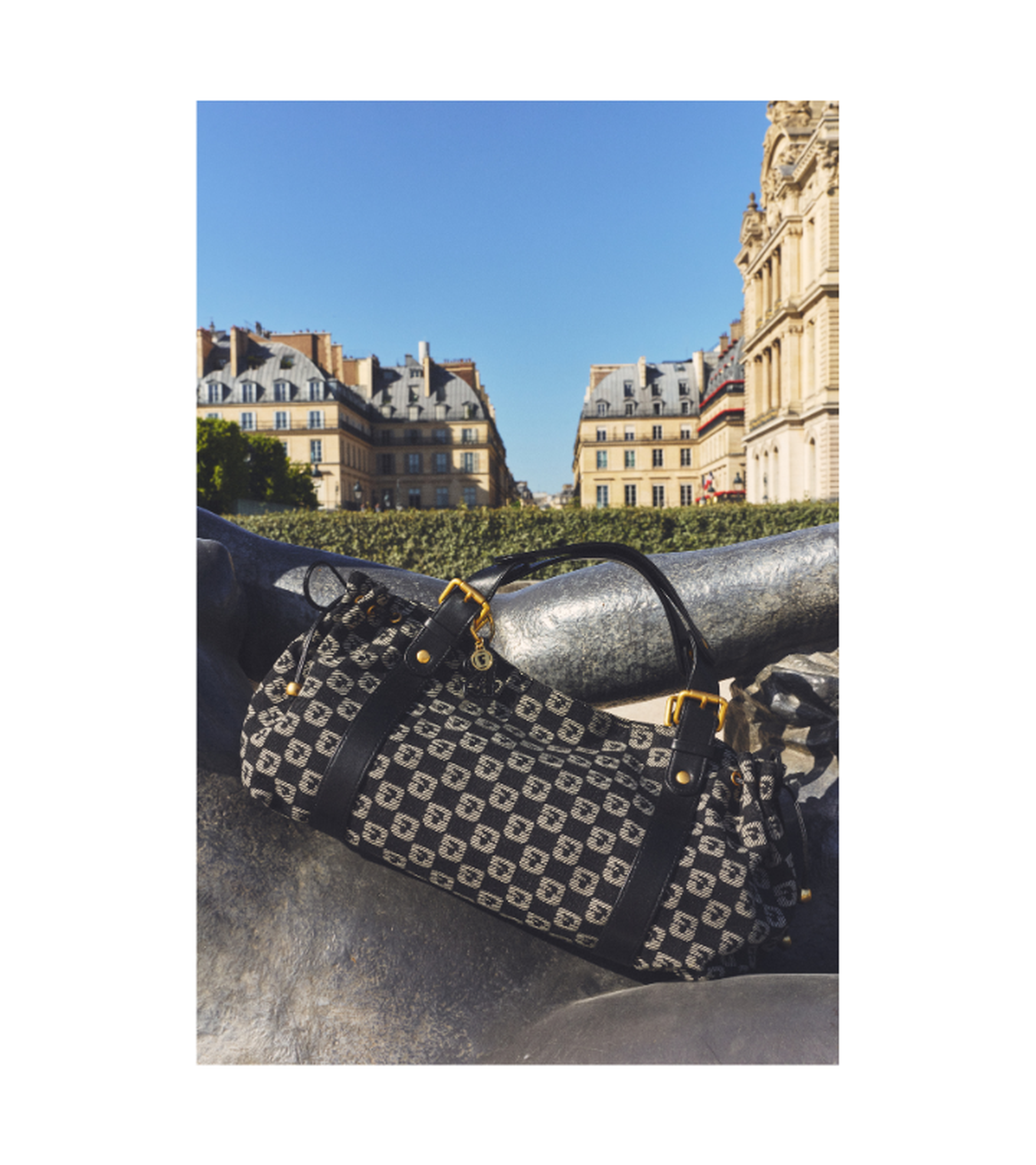 Patterned handbag rests on a statue with historic European buildings in the background, under a clear blue sky.