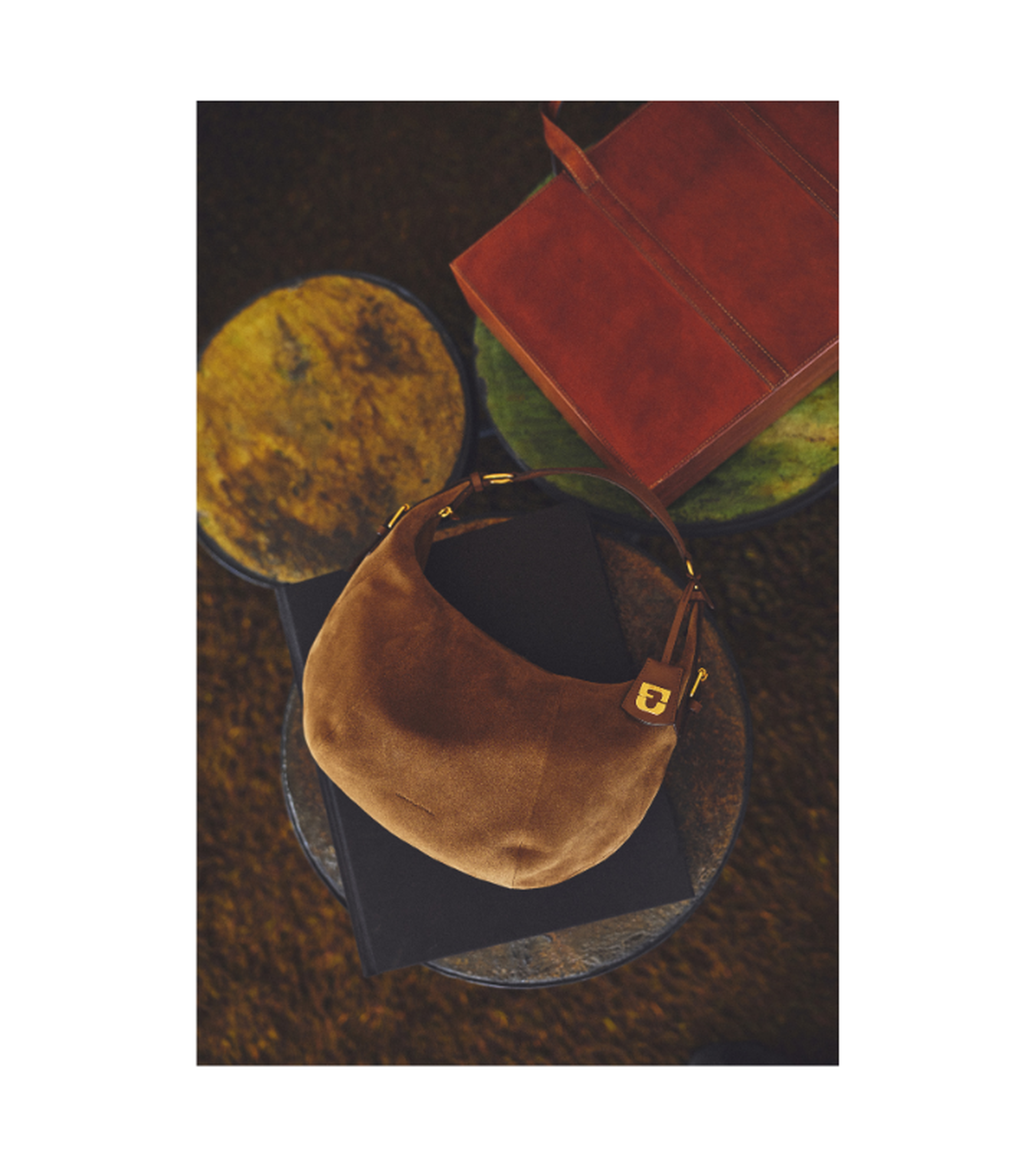 A brown suede handbag with a curved shape, placed on a round table alongside a red leather wallet and a black book.